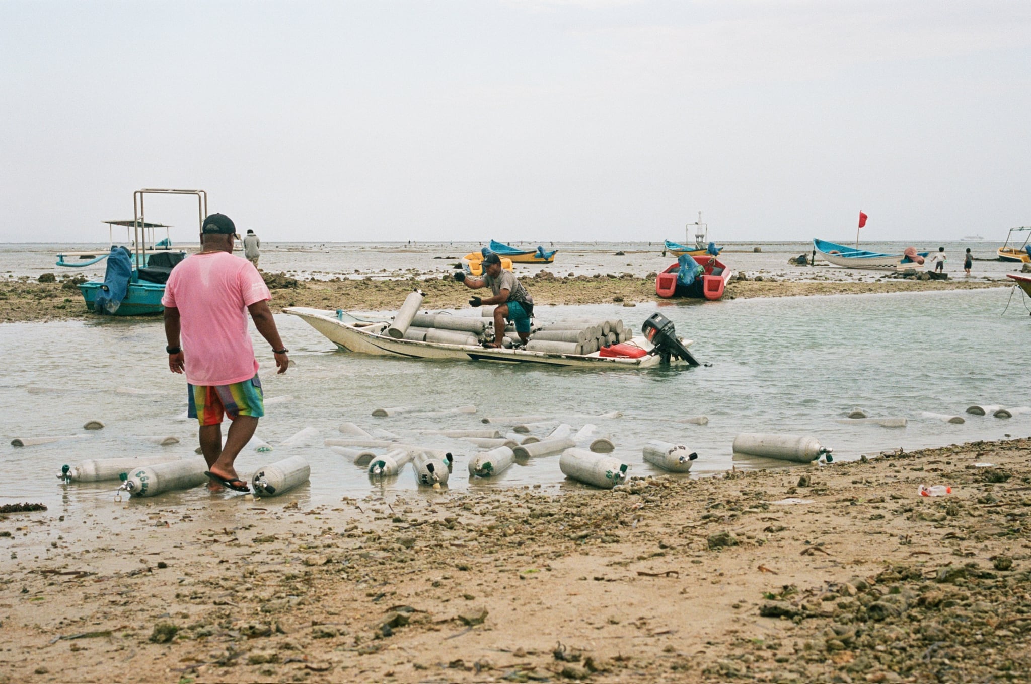 A person in a pink shirt wades toward small boats in shallow coastal water.