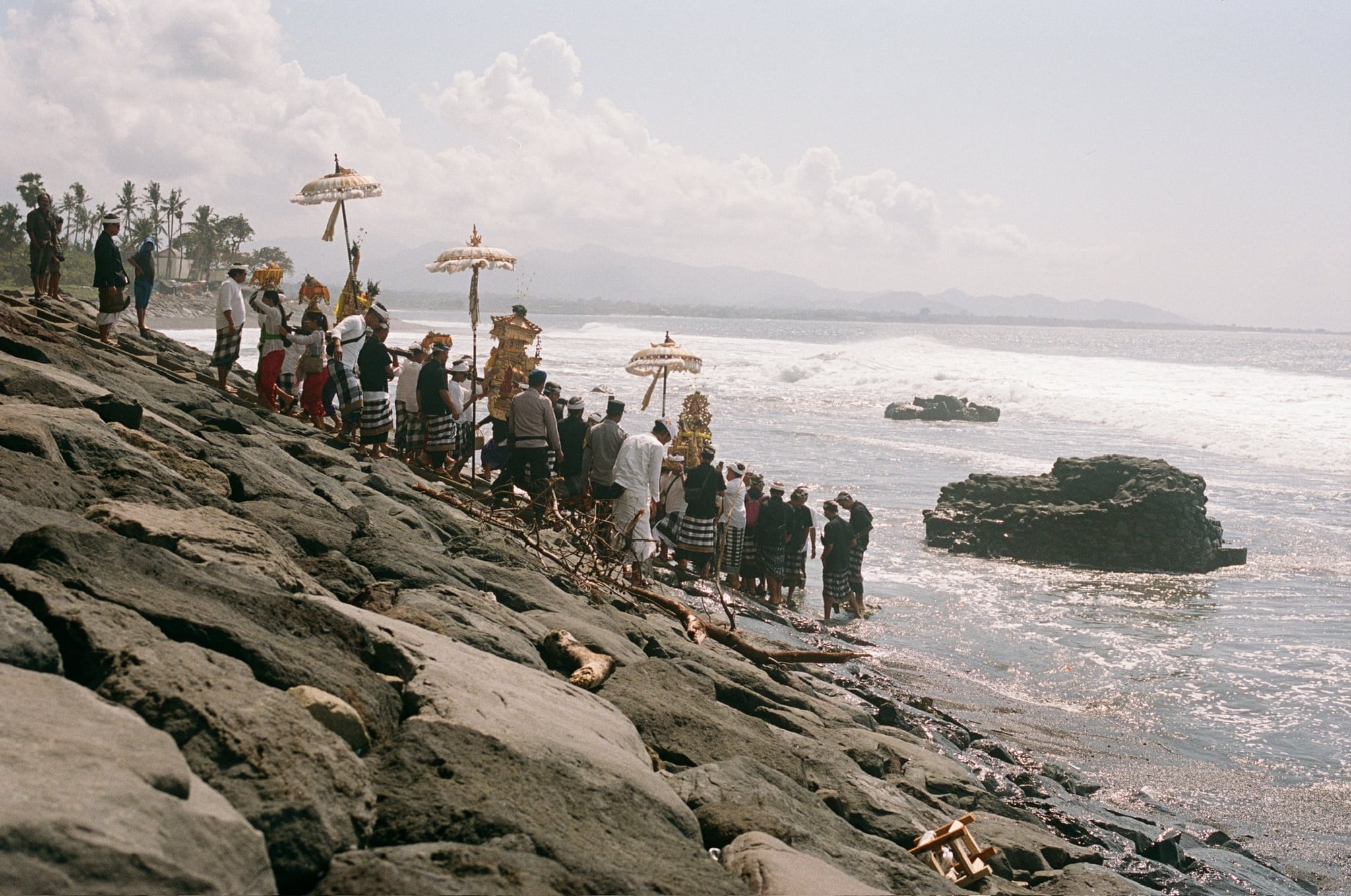 A line of people carrying tall umbrellas stands along a sloping stone sea wall above the surf.