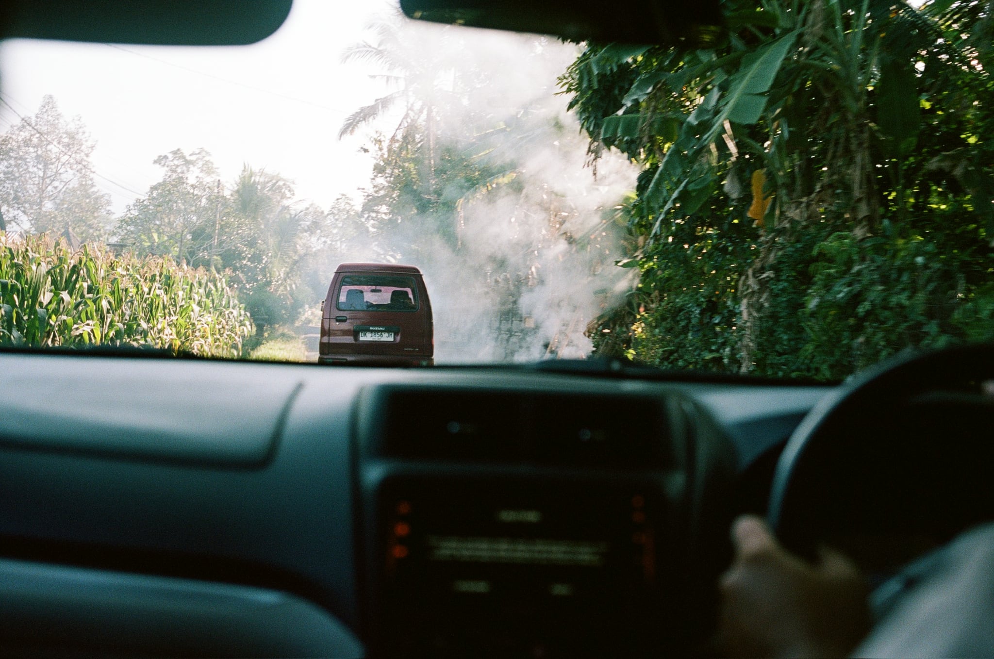 A car ahead is seen through a vehicle windscreen on a narrow road bordered by greenery.