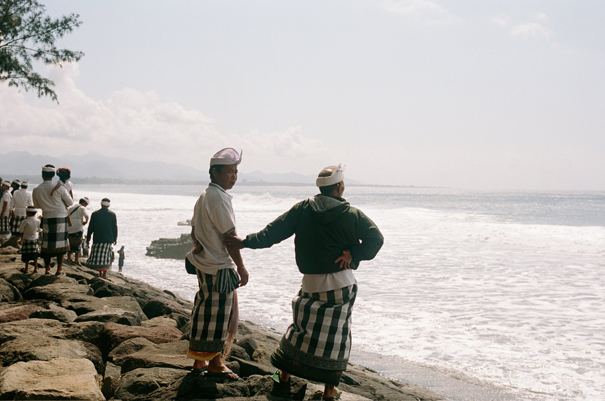 People in white headwear stand on a stone embankment looking across the sea.