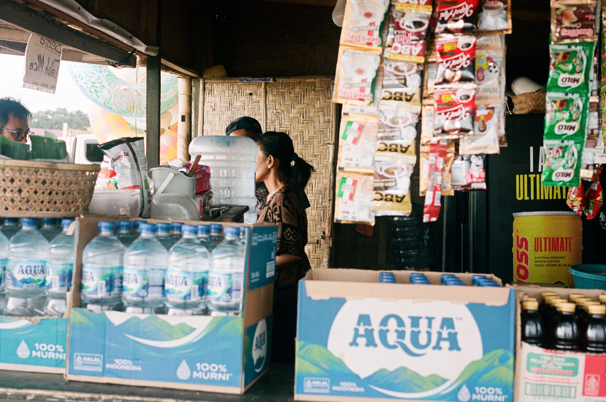 Bottled water and hanging packets crowd the counter of a small shaded shop.