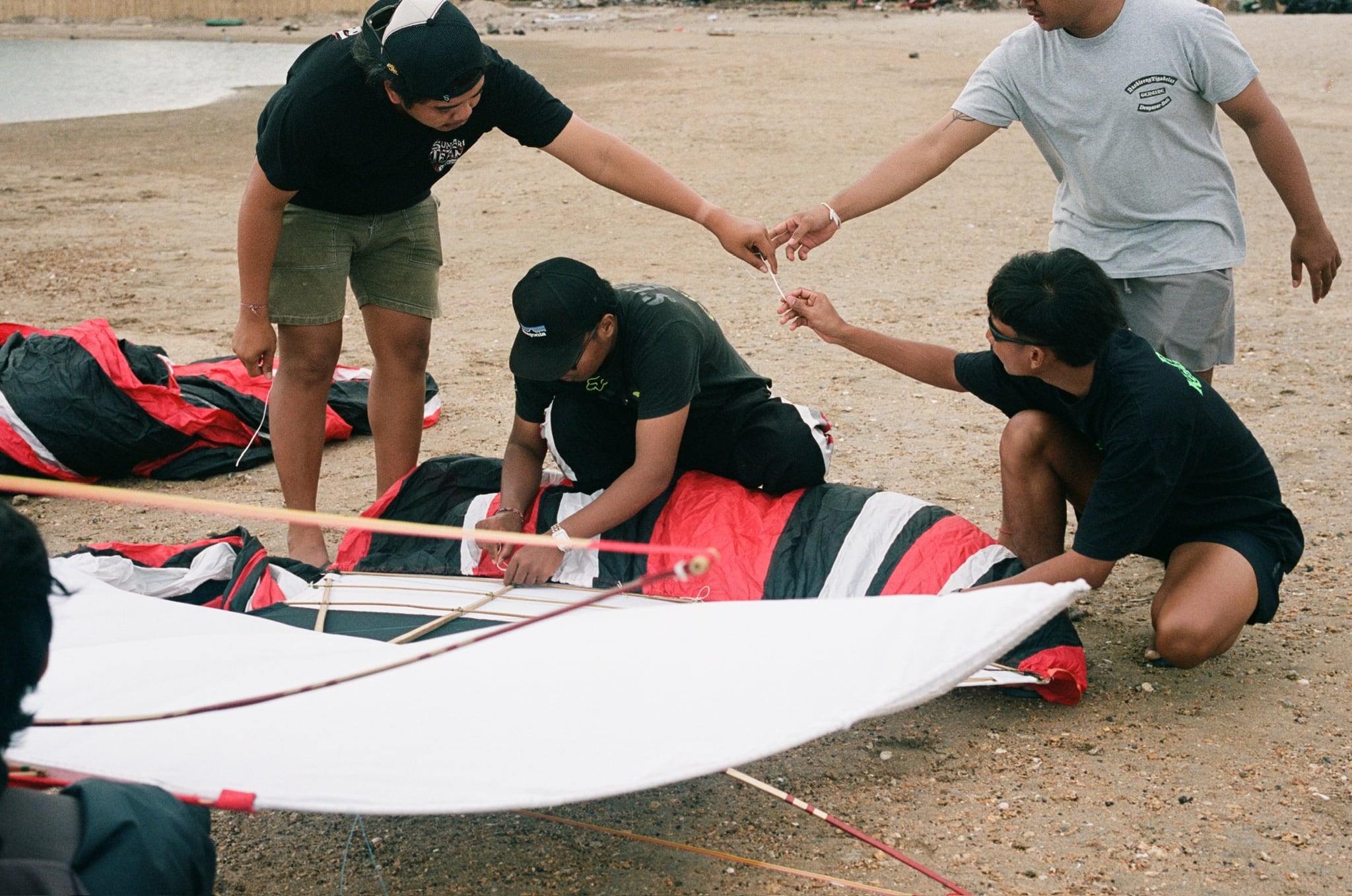 A group crouches around a large white kite with red-and-black fabric on sandy ground.