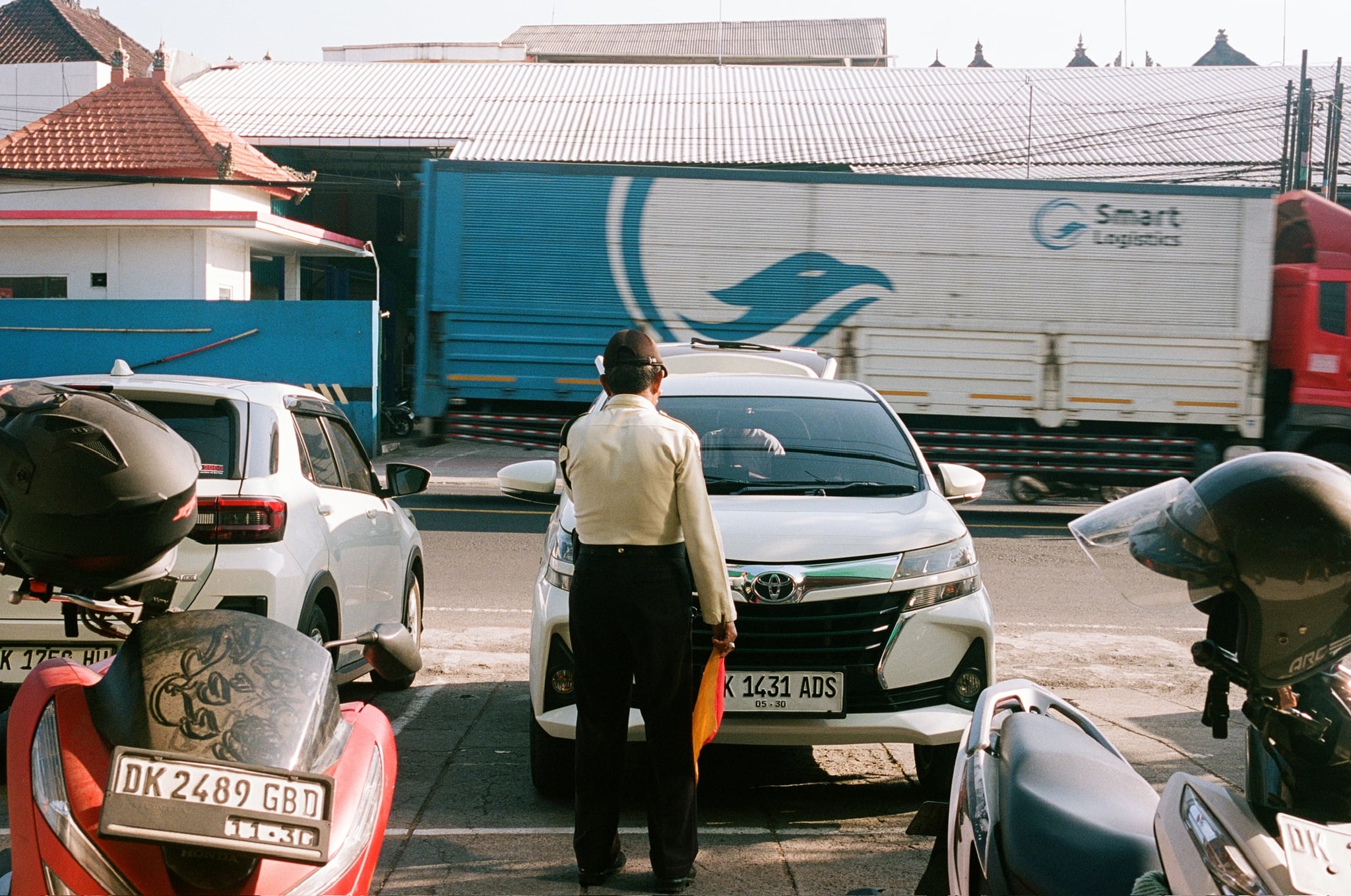 A person stands between parked vehicles while a large truck passes on the road behind.