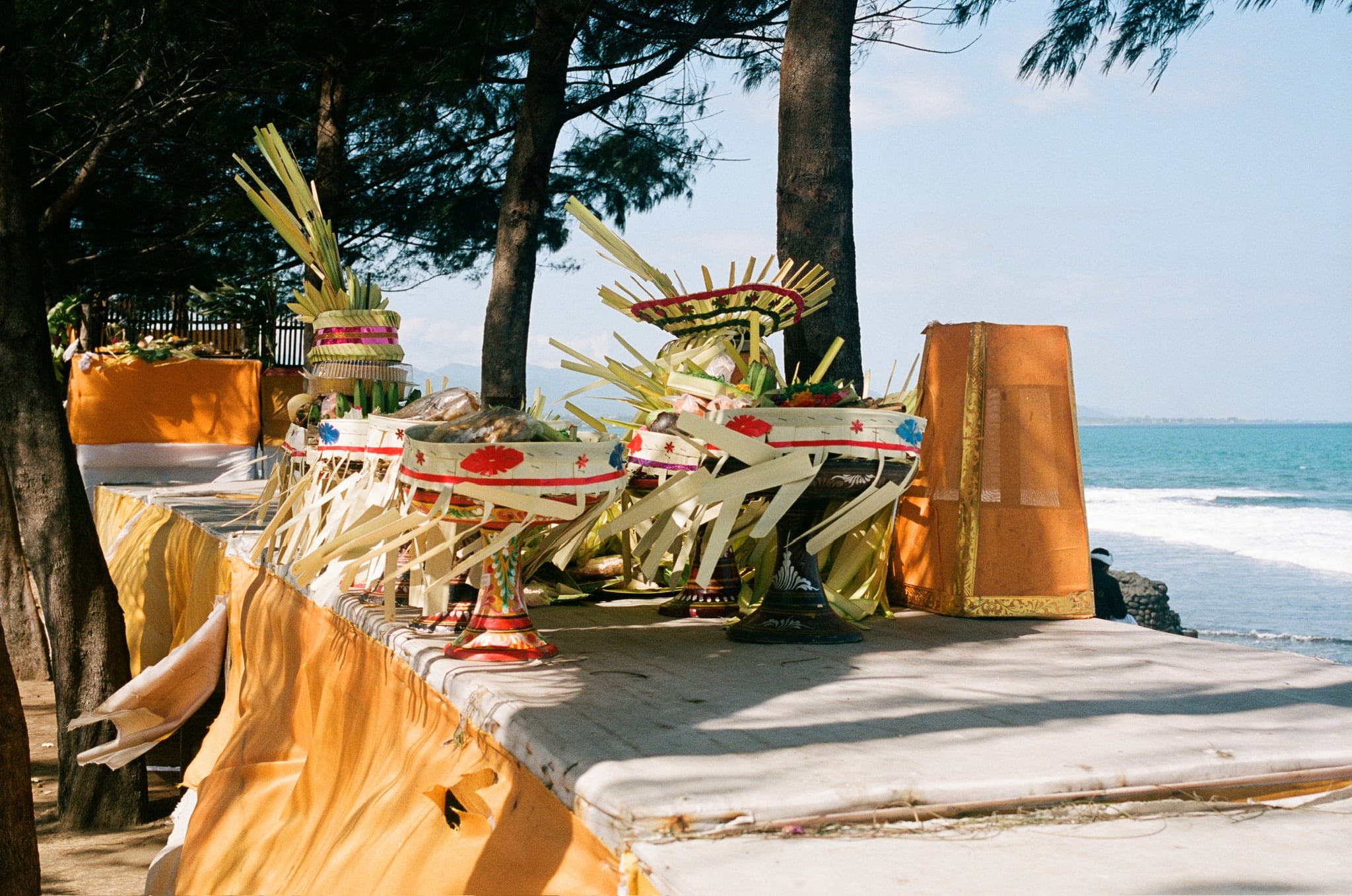 Woven leaf arrangements and containers sit on a yellow-draped table beneath trees beside the sea.