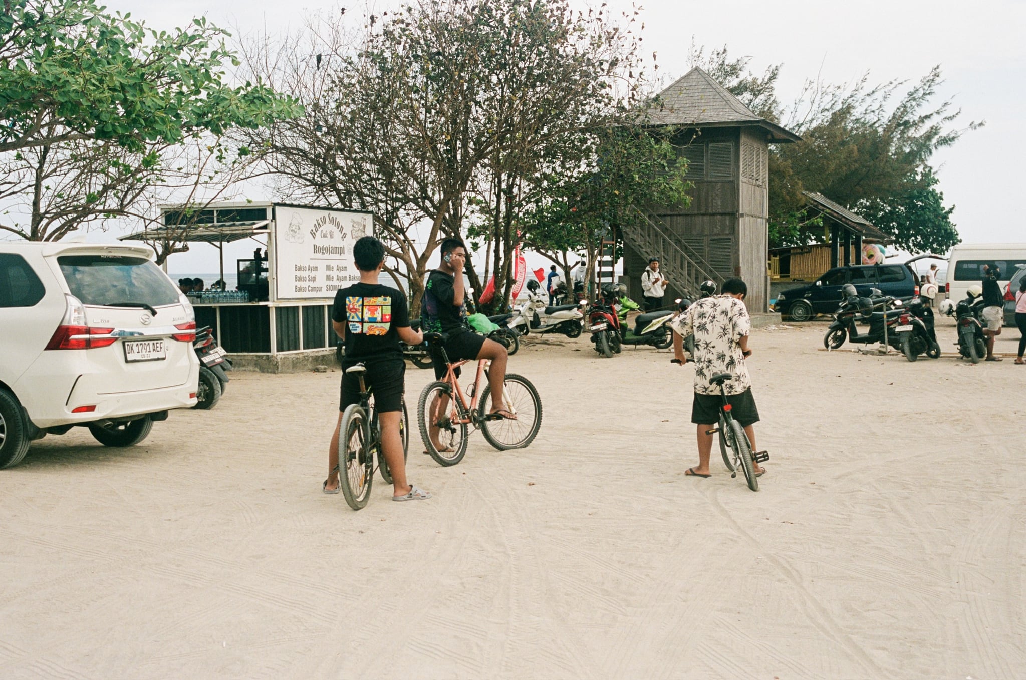 People with bicycles pause in a broad open lot bordered by vehicles and low buildings.