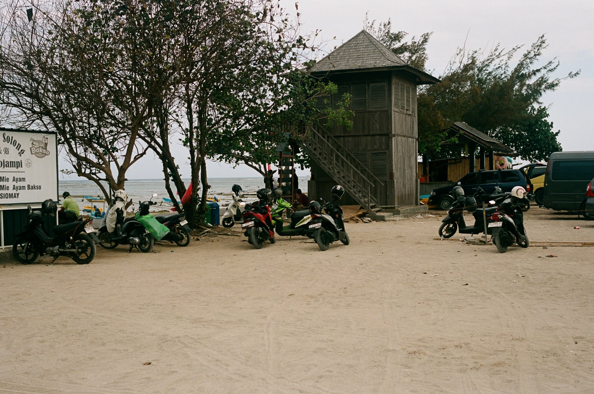 Scooters and people gather beside a tall gate and trees at the edge of an open lot.