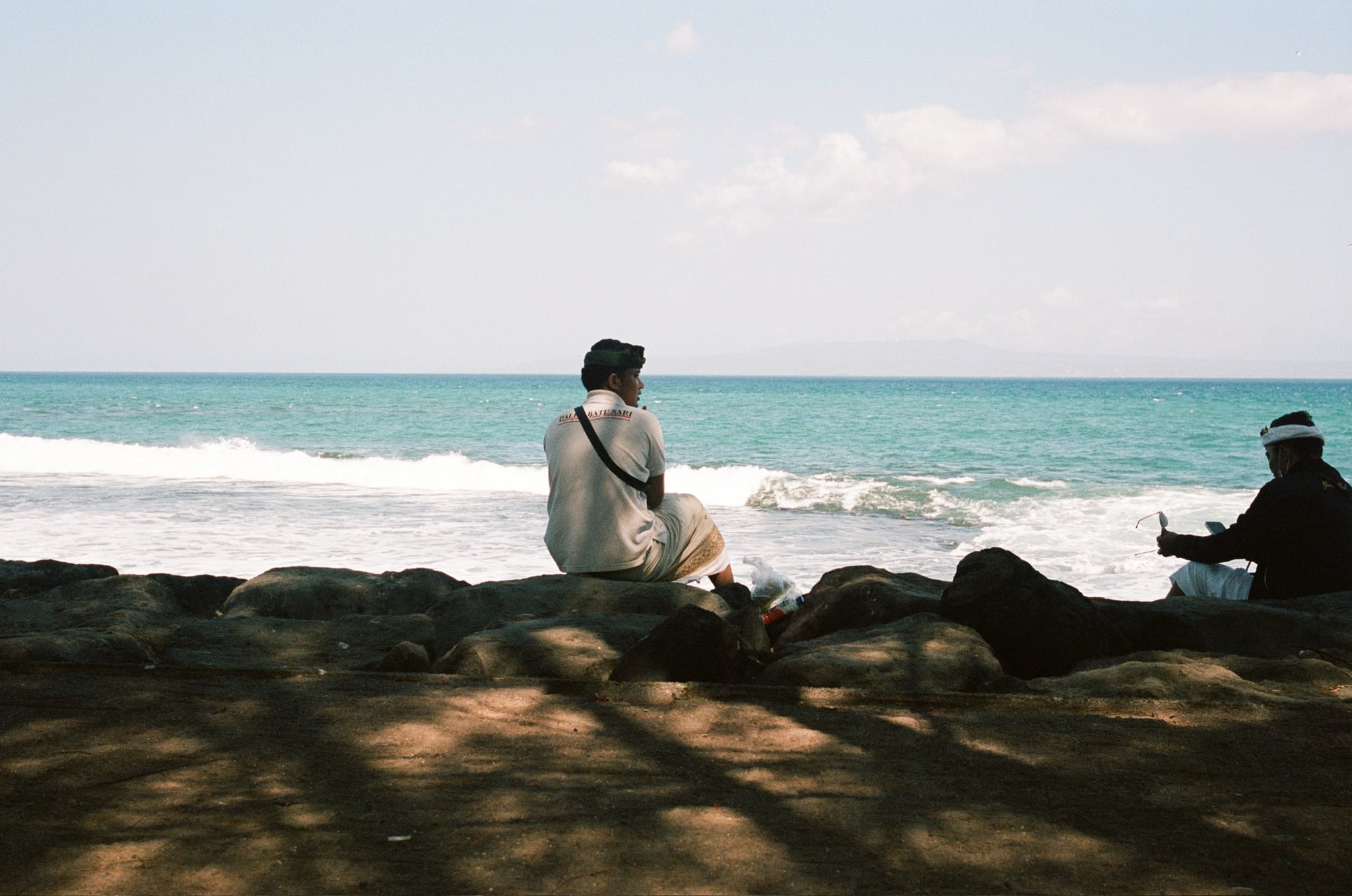 Two people sit separately on rocks in the shade, facing turquoise water and white surf.