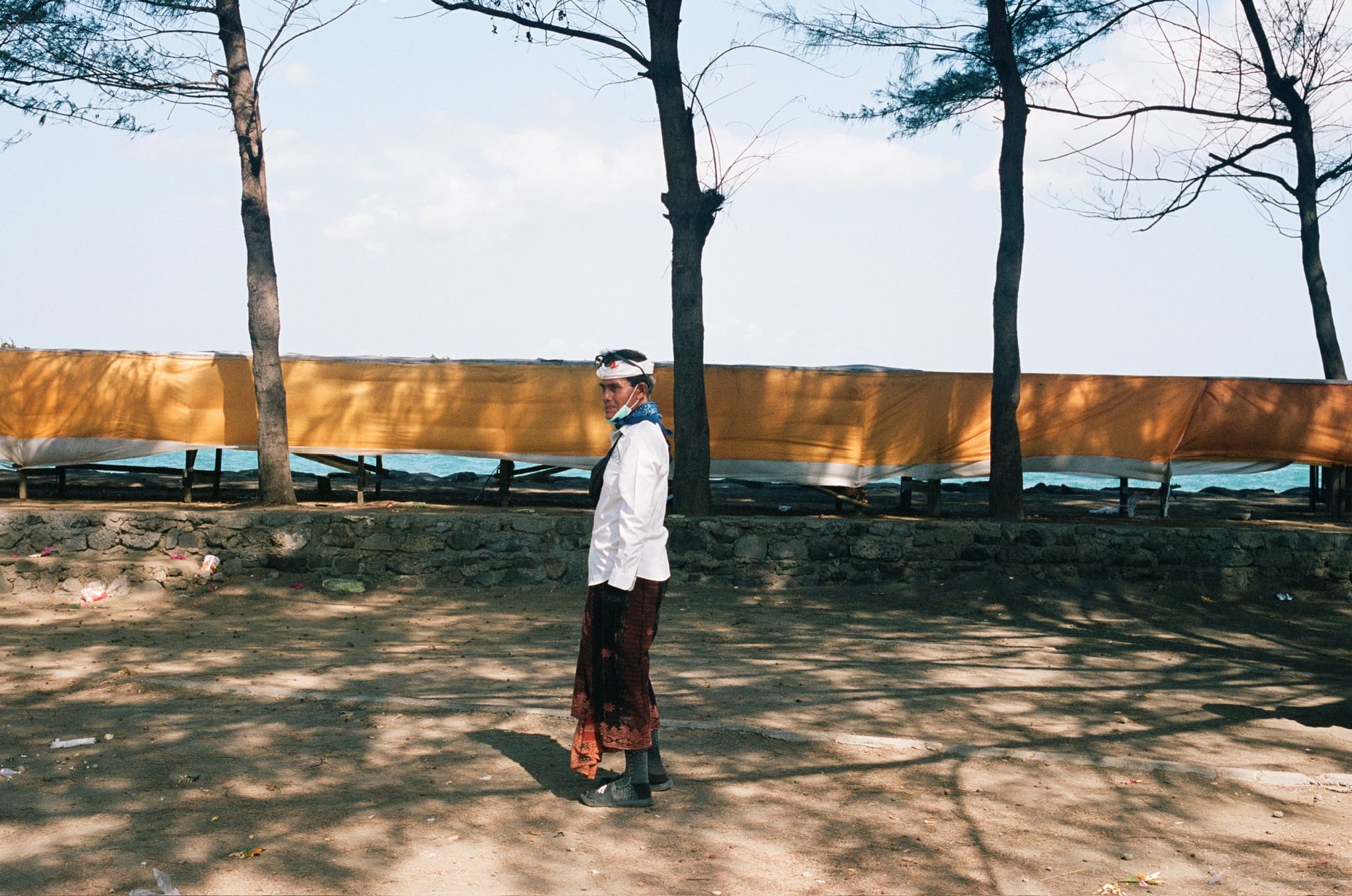 A person in white clothing walks through tree shadows beside a yellow fabric screen and the sea.