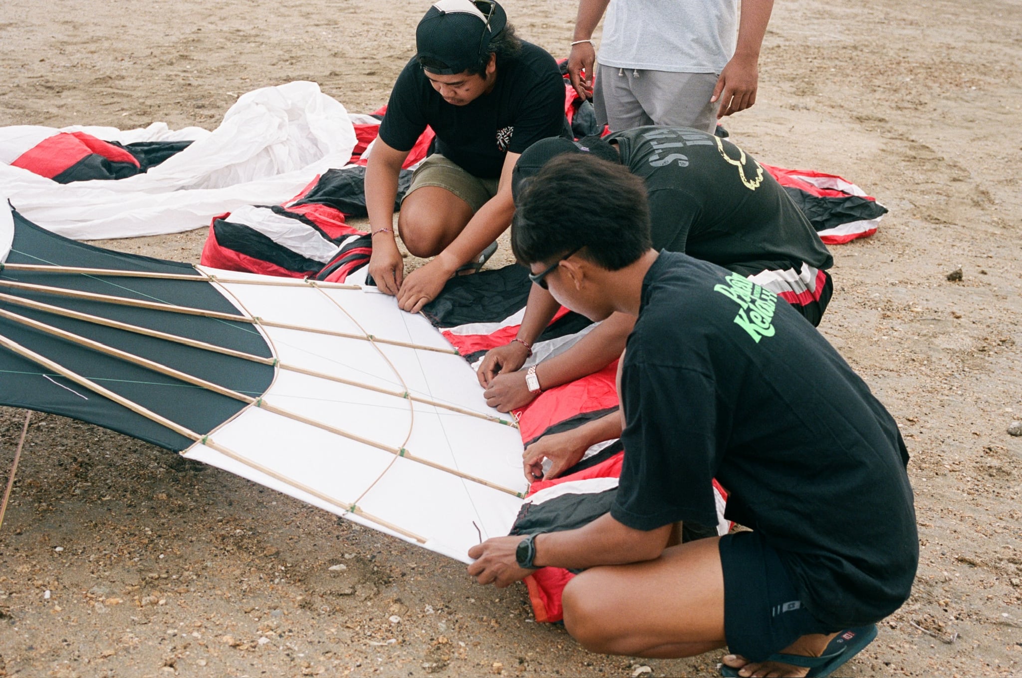 People stretch white fabric over the curved frame of a large kite on sandy ground.