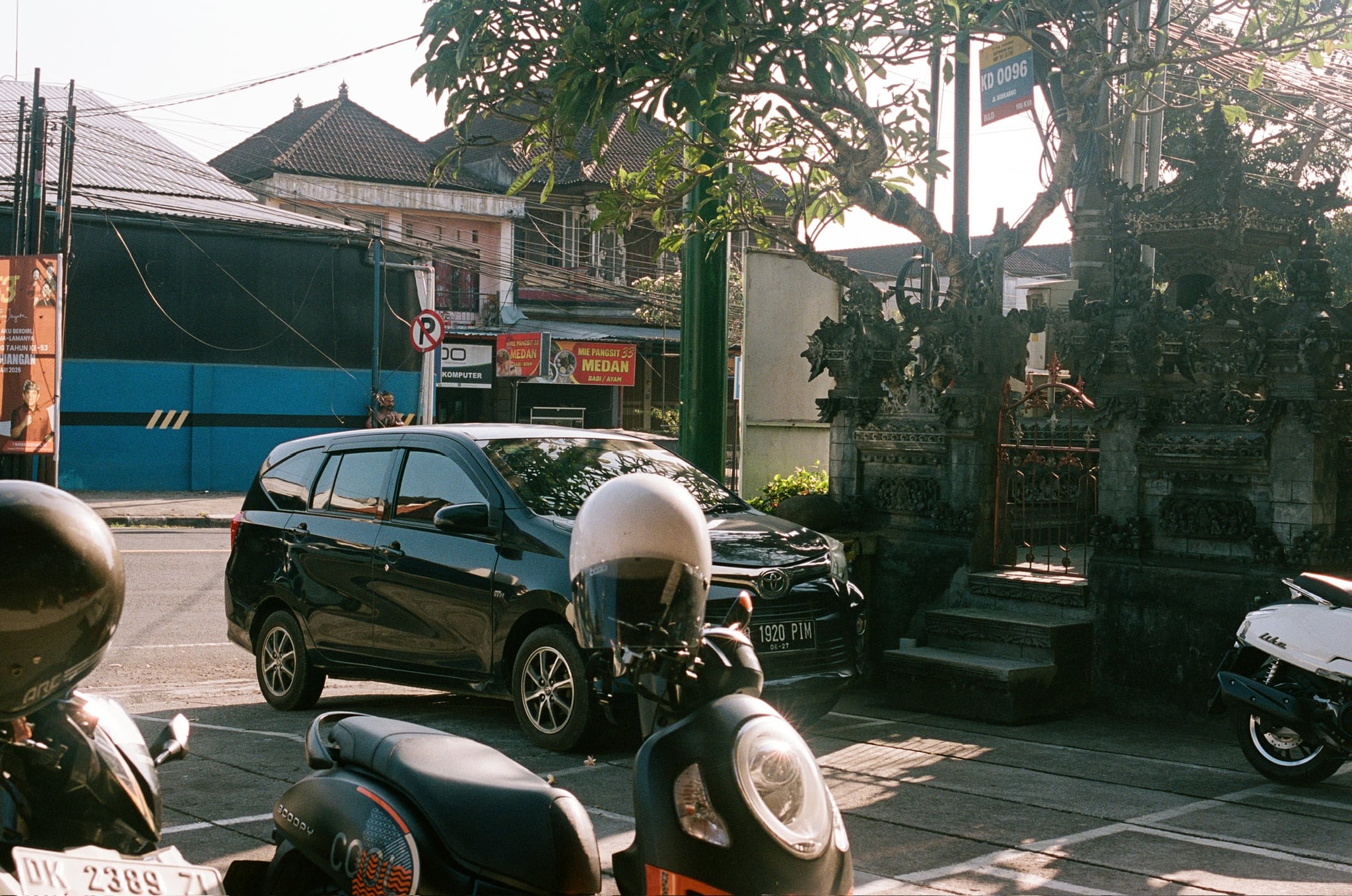 Scooters and a dark car stand along a sunlit street with low shopfronts behind.