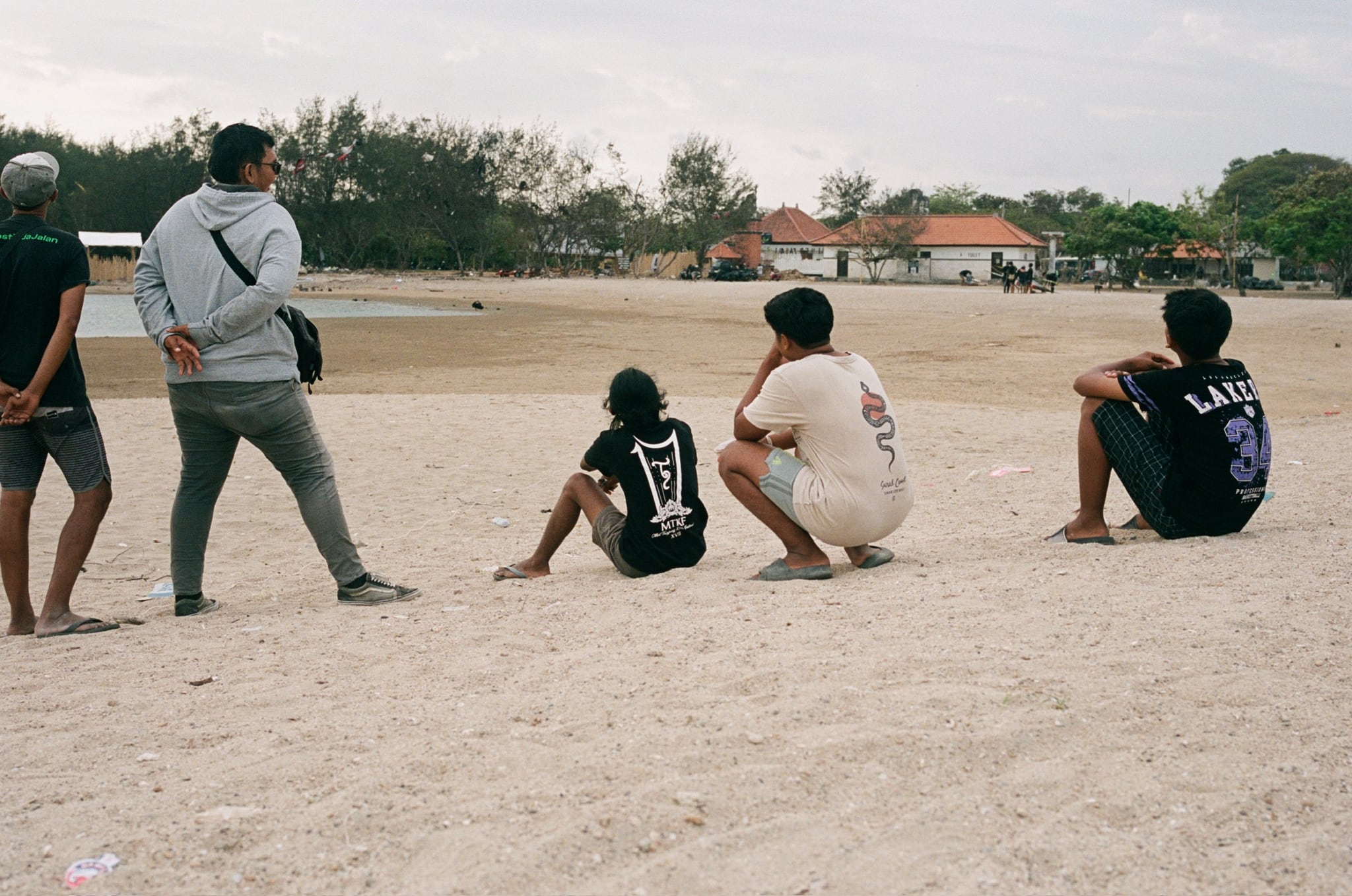 People sit and stand on pale open ground beneath a softly clouded sky.