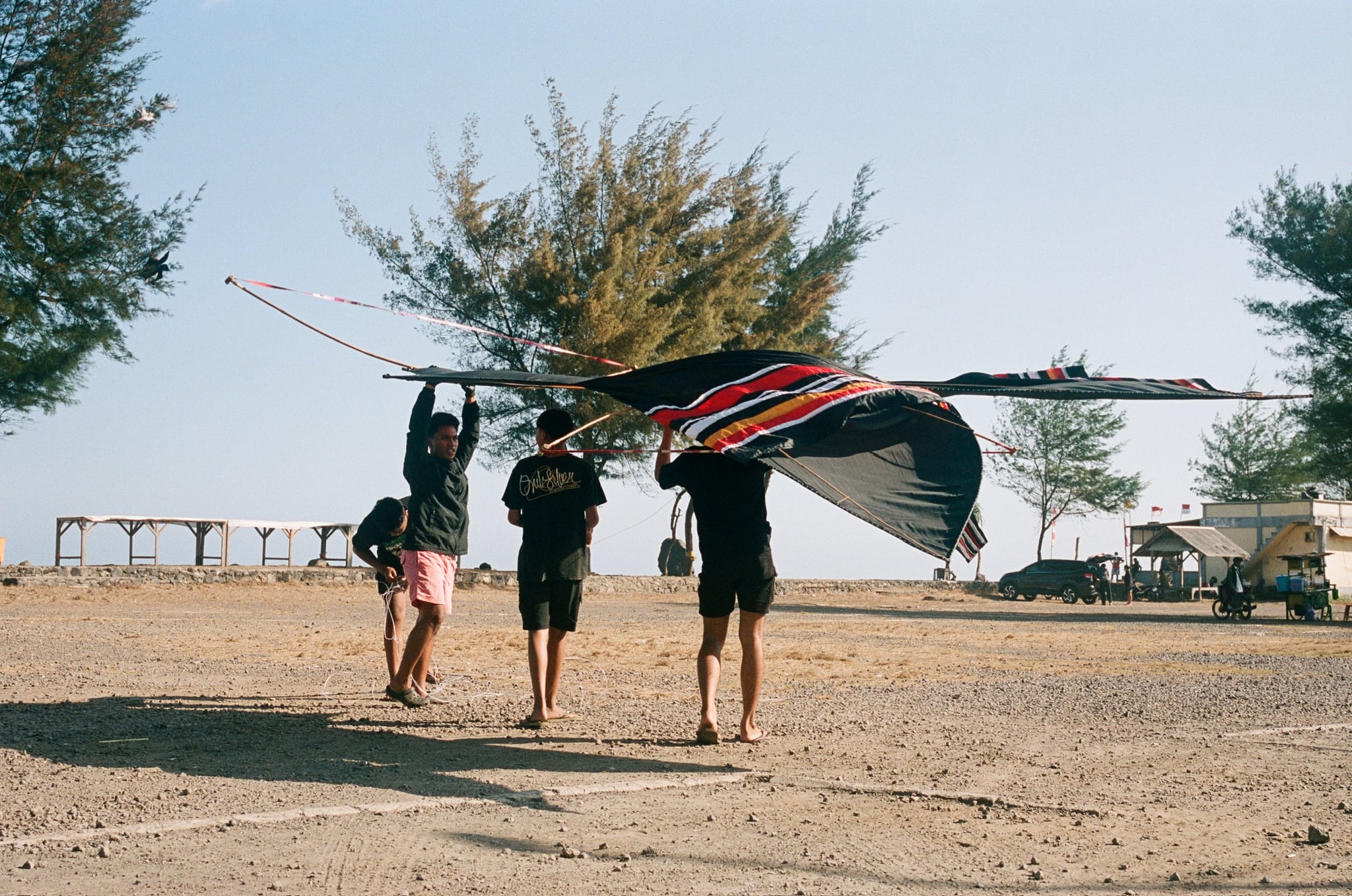 Three people carry a large, colourful kite overhead across open ground near the coast.