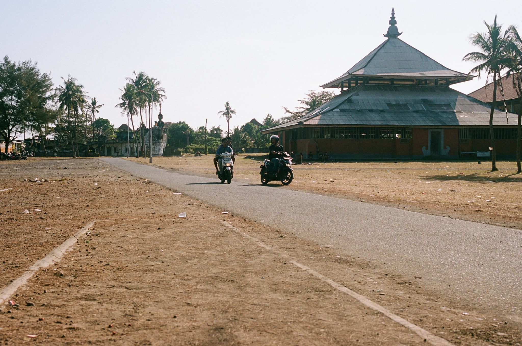 Motorcycles pass a large tiered-roof pavilion beside a wide, dry field.