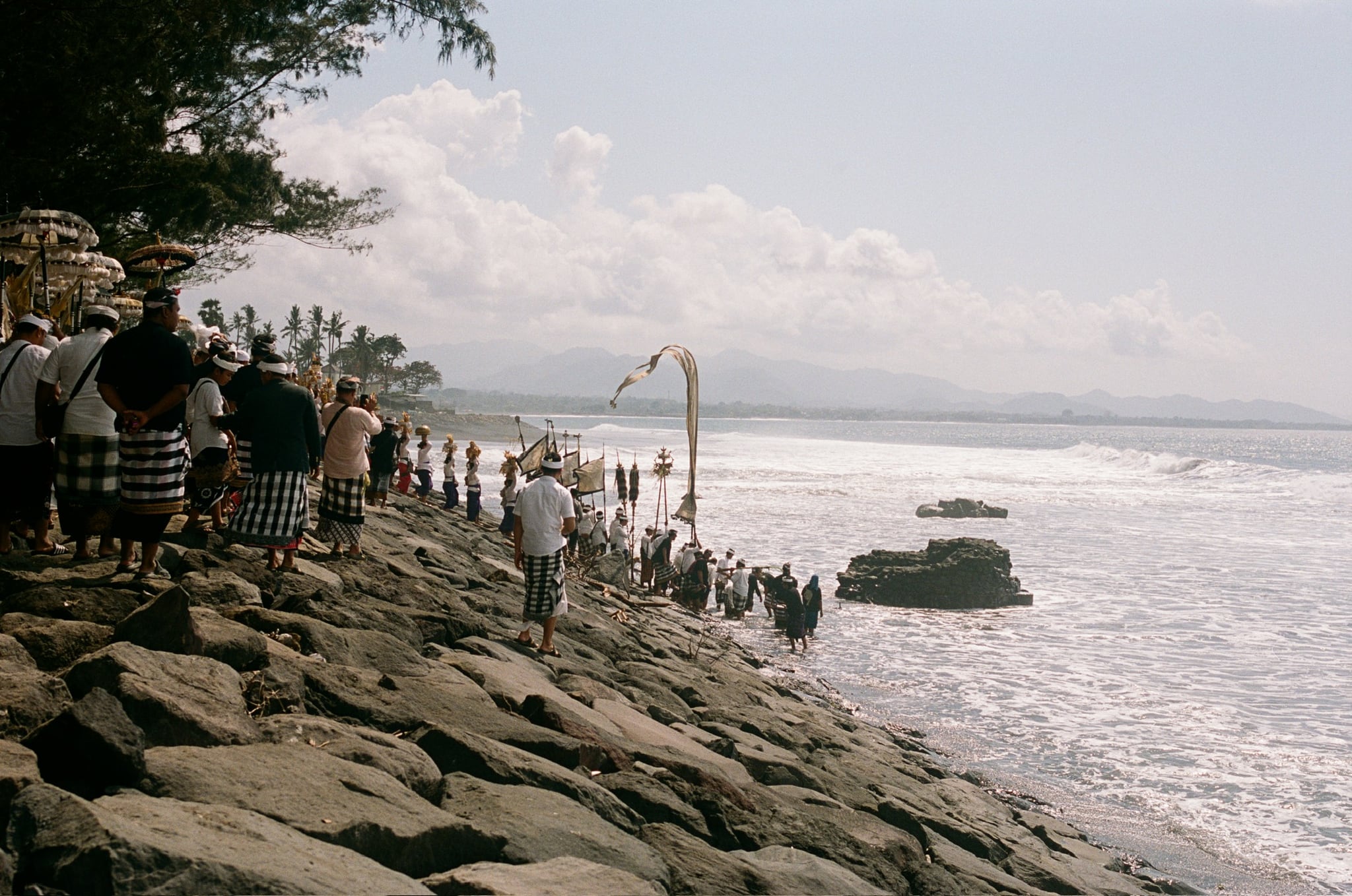 People stand in a long line on a stone coastal embankment, with tall umbrellas above them.