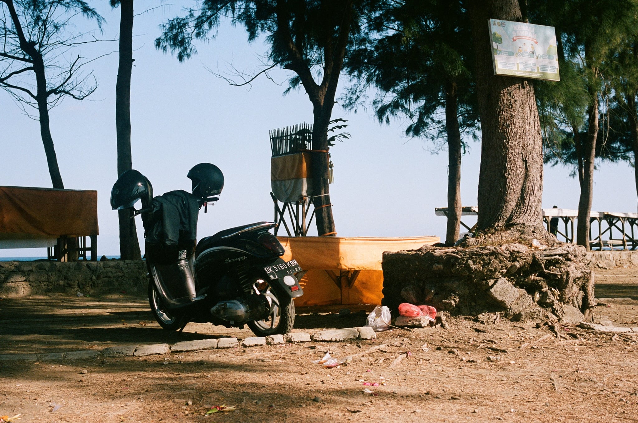 A scooter rests beneath trees beside a yellow screen, with the sea visible beyond.