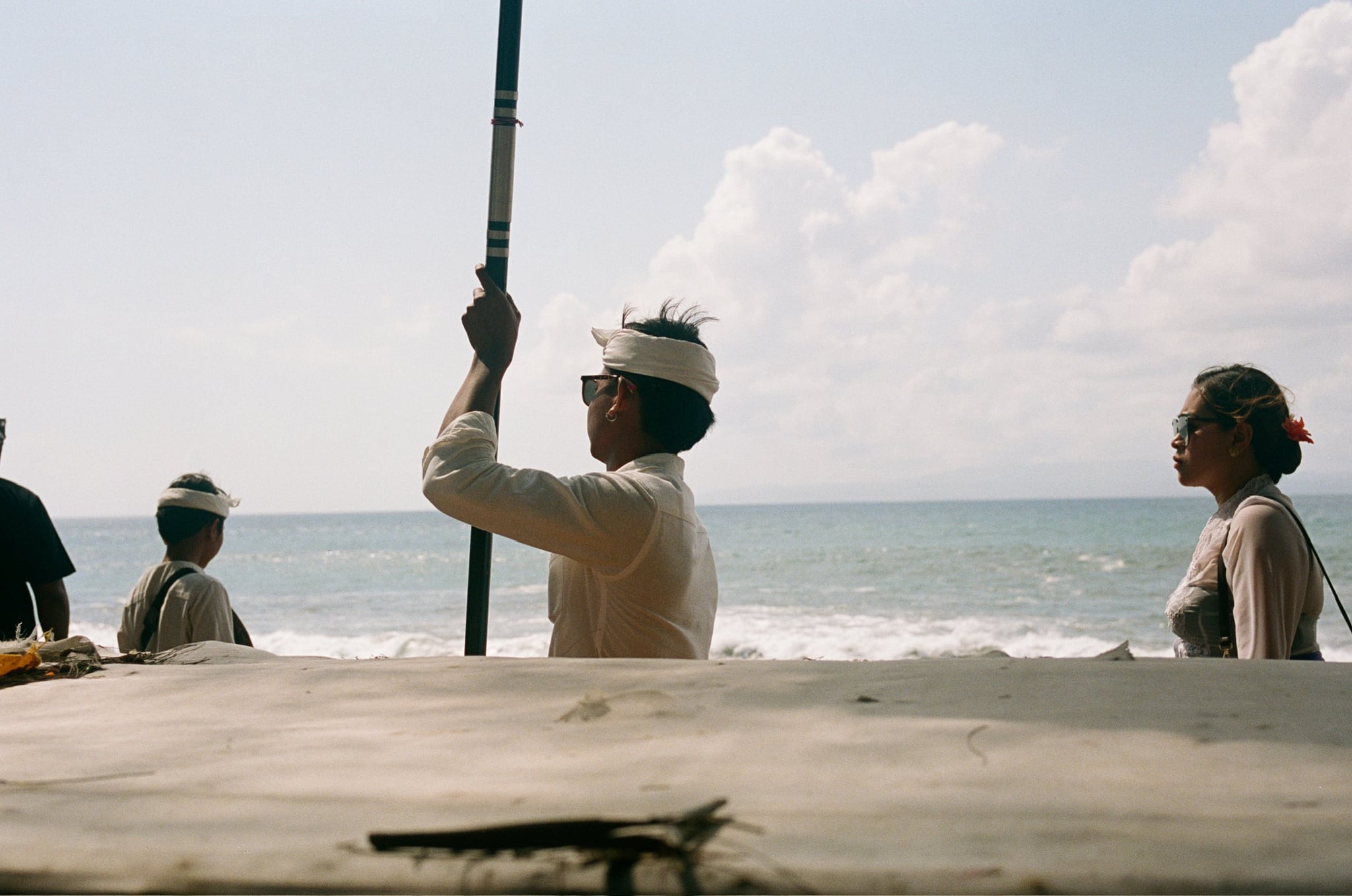 A person in white headwear holds a tall pole beside the sea, with another person nearby.