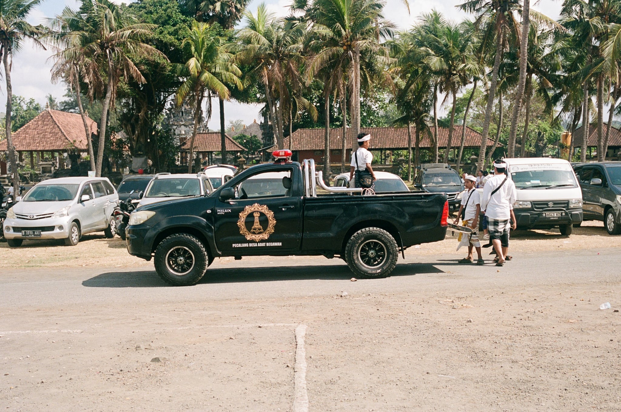 A dark pickup truck moves past parked cars and people beneath tall palms.