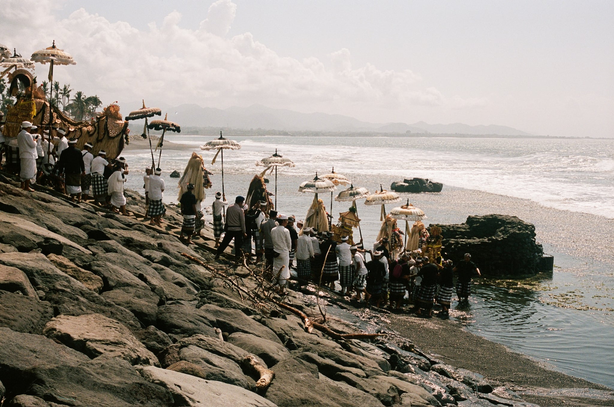 Tall umbrellas and people form a line down a stone embankment toward the surf.
