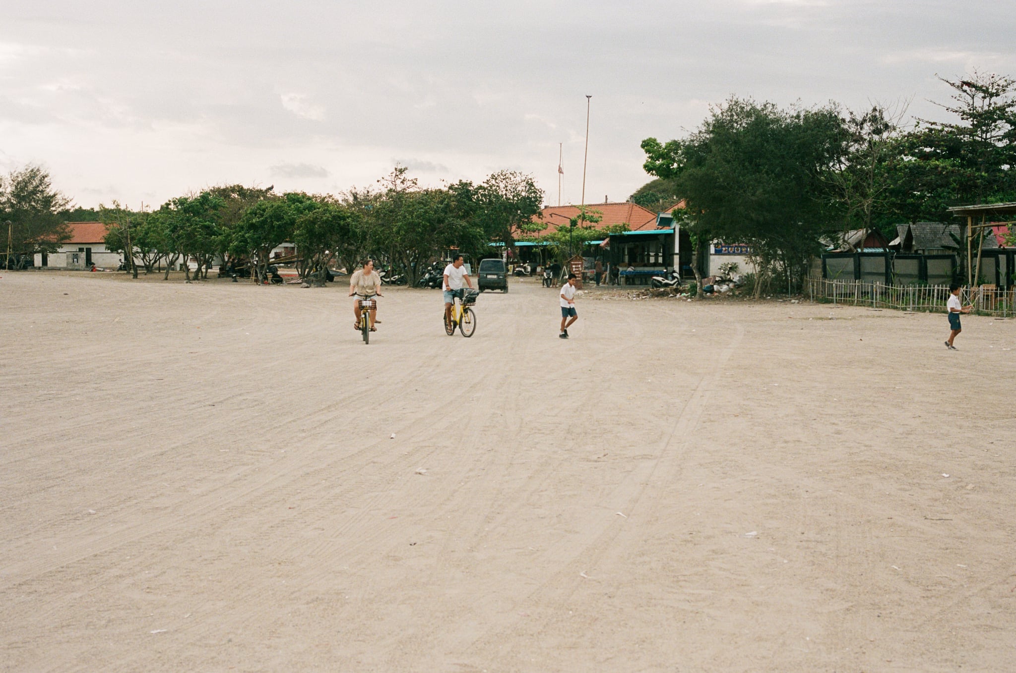 Scattered figures cross a wide, pale field bordered by trees and low buildings.