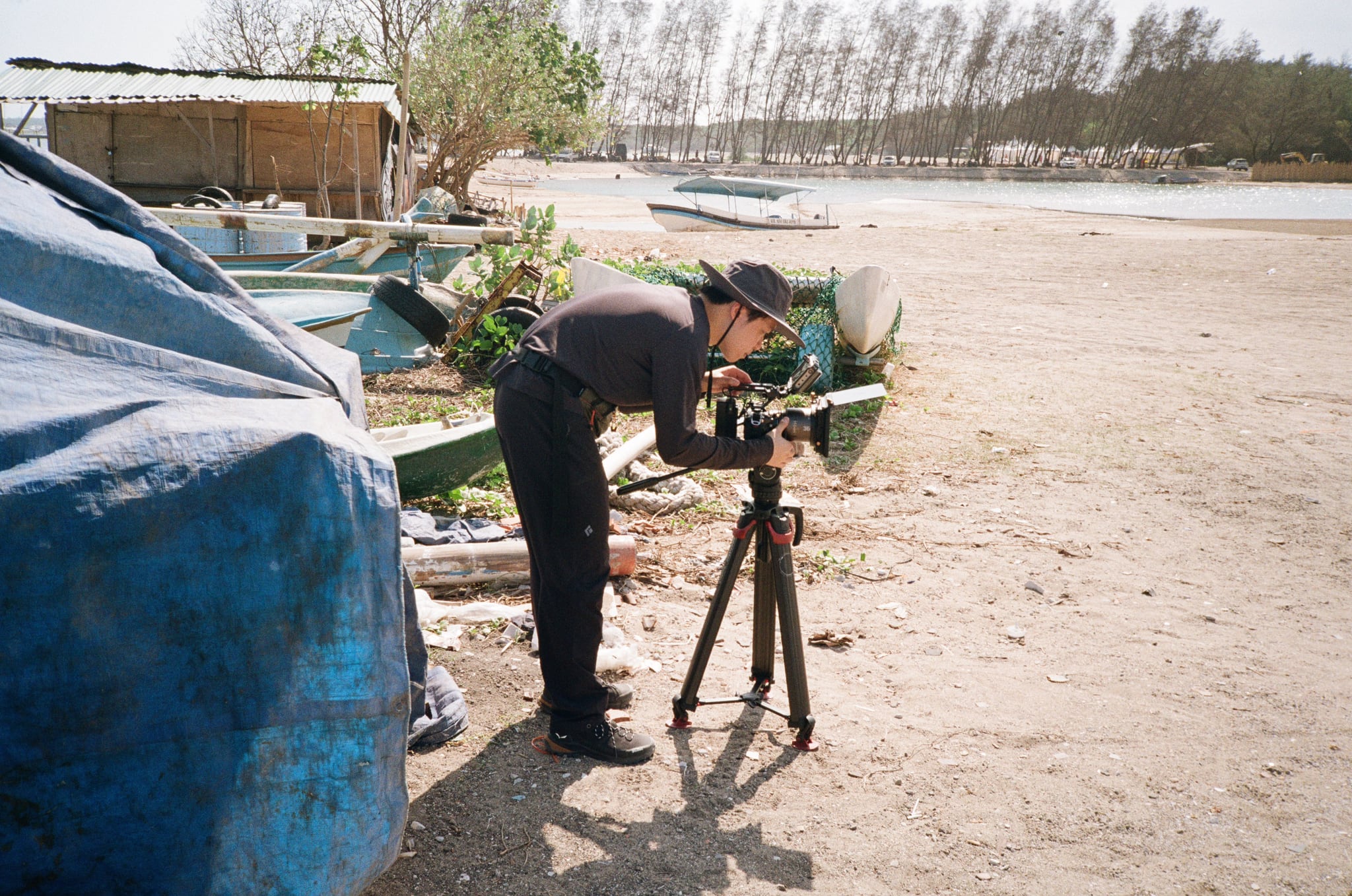 A person bends beside a tripod and covered boats on sandy coastal ground.