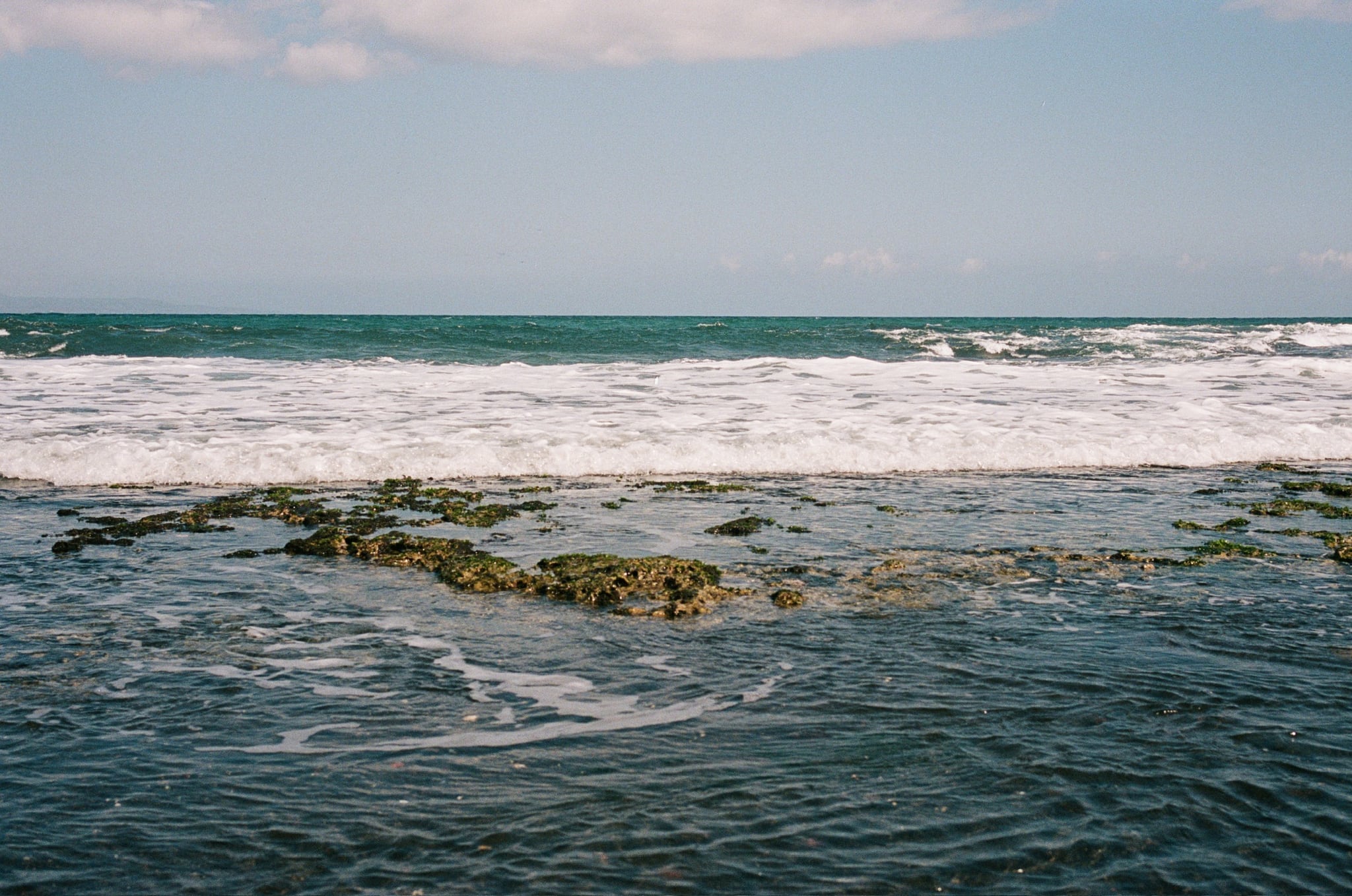 White waves roll across a shallow rocky reef beneath a pale blue sky.