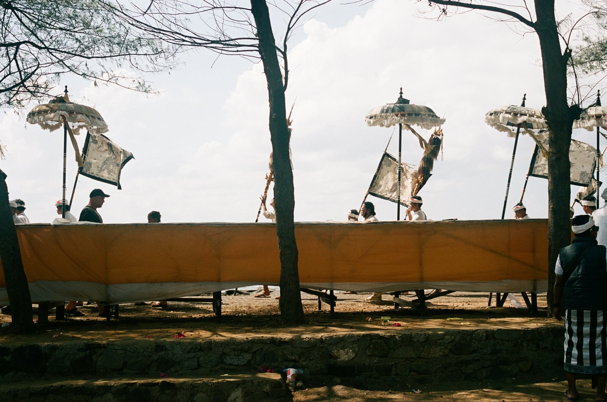 Tall decorated banners rise between trees behind a yellow fabric screen beside the sea.
