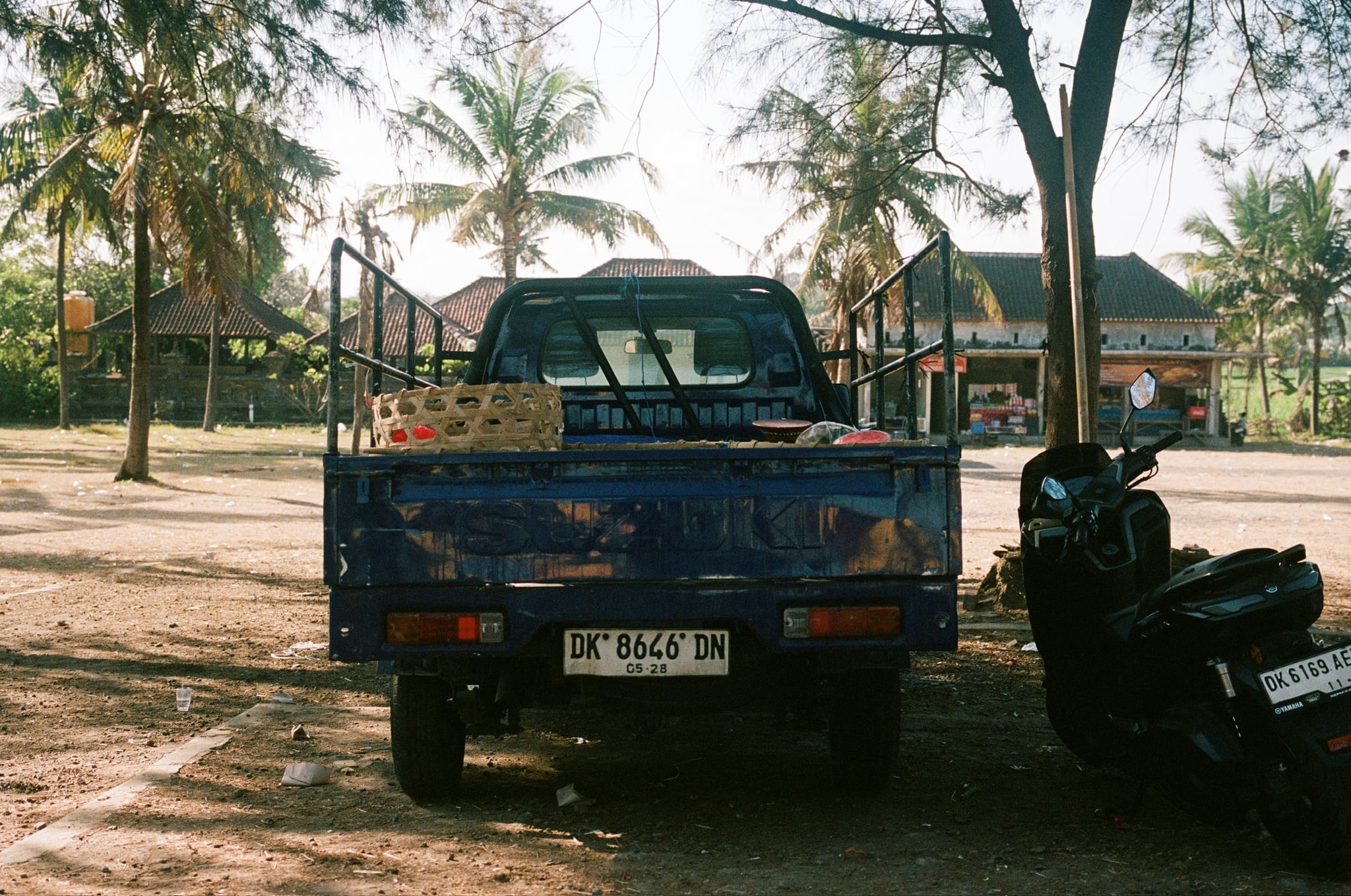 A blue pickup and a scooter stand beneath palm trees beside low buildings.