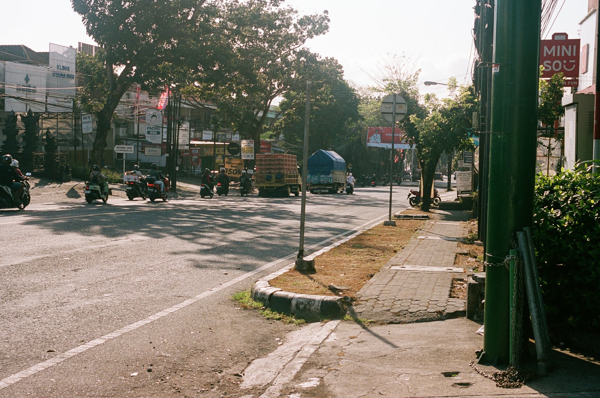 Low sunlight crosses a city junction with road markings, trees, and roadside buildings.