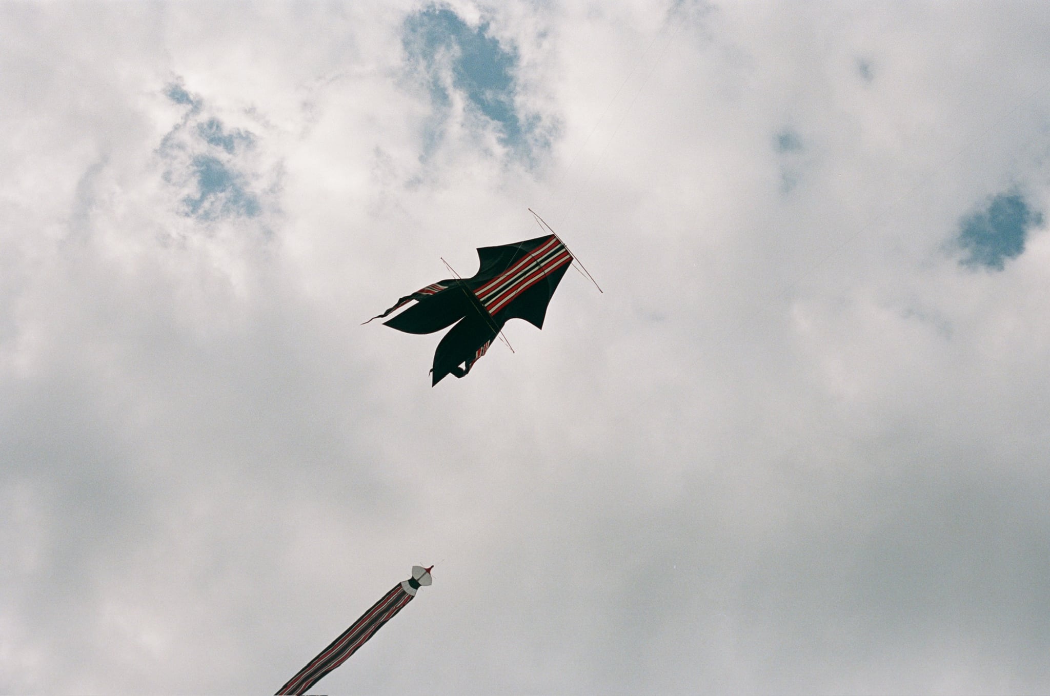 A red, green, and dark kite flies above long kite tails against a cloud-filled sky.
