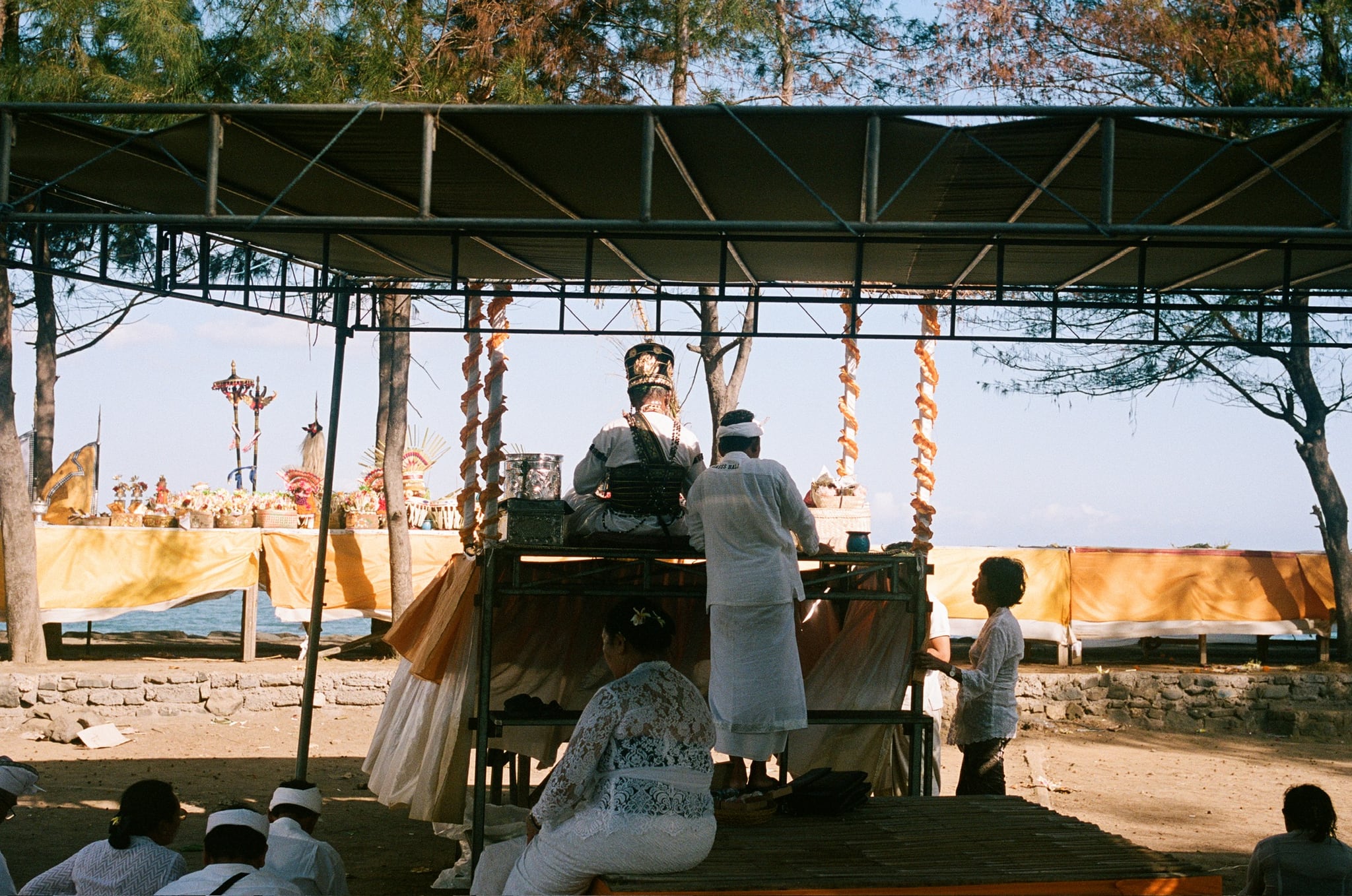 People gather around a decorated platform beneath a broad open-sided roof.