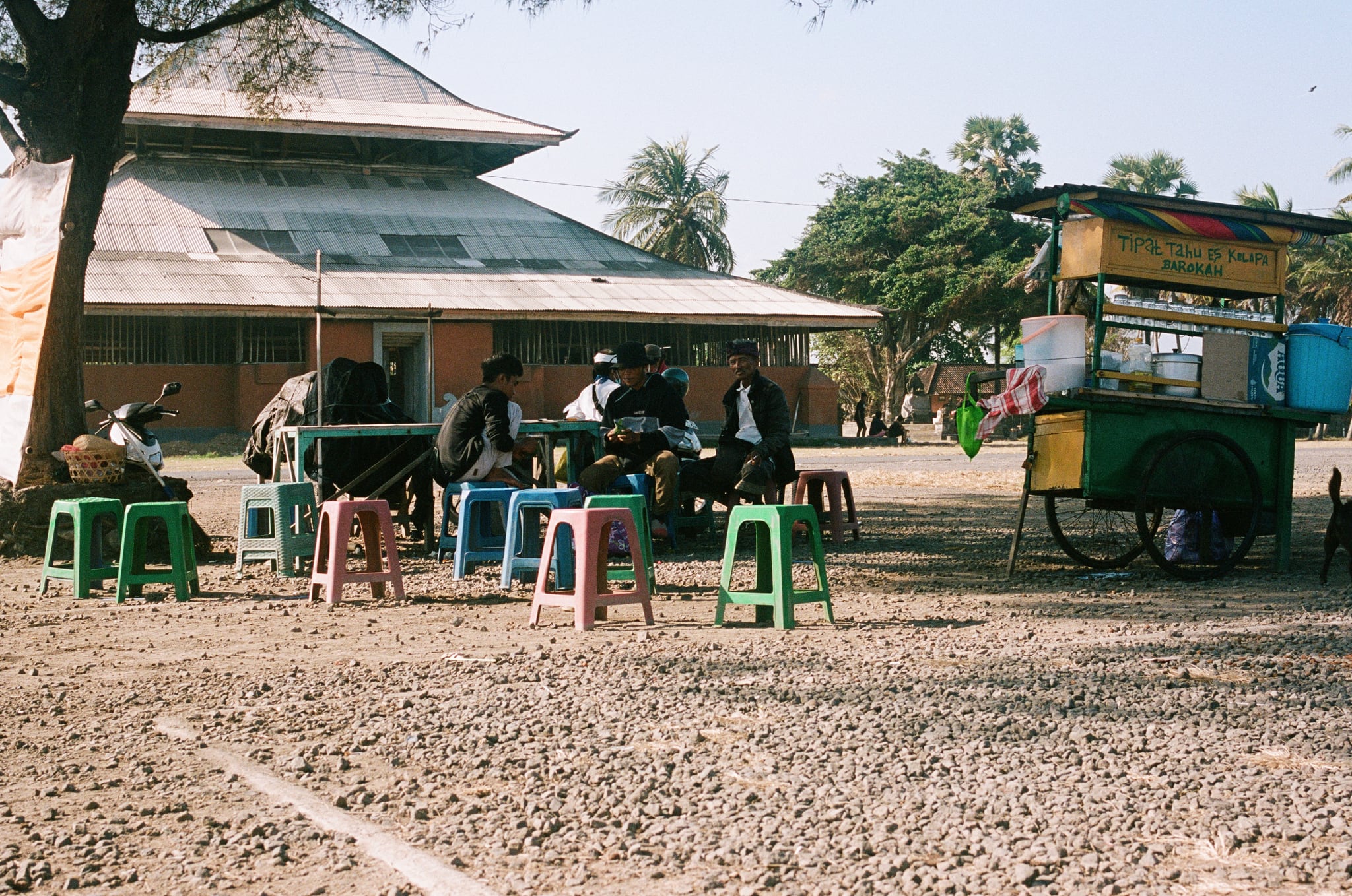 People sit on colourful plastic stools beside a roadside cart and a large pavilion.