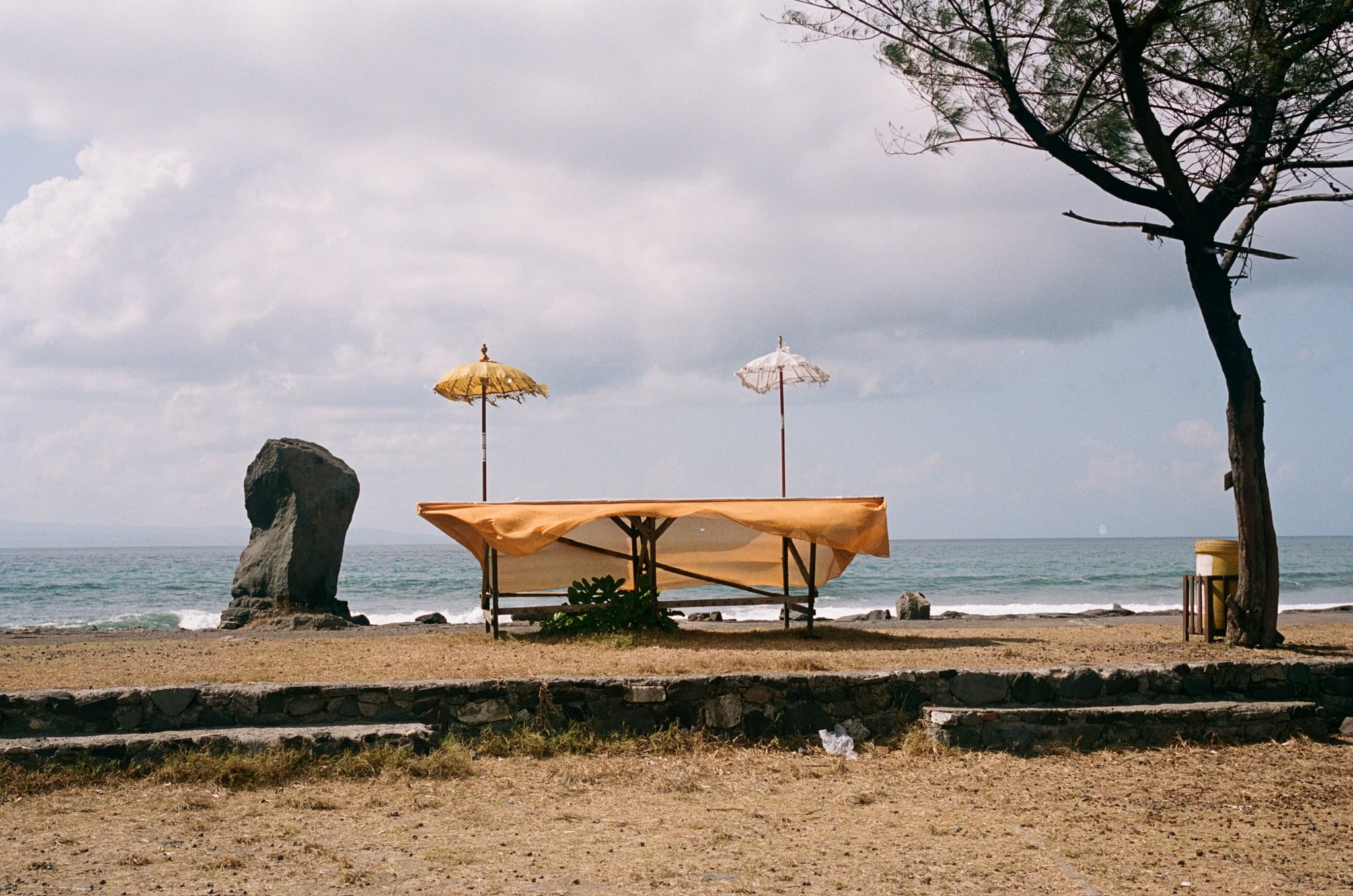 An empty yellow-draped canopy and two umbrellas stand beside the sea beneath a cloudy sky.