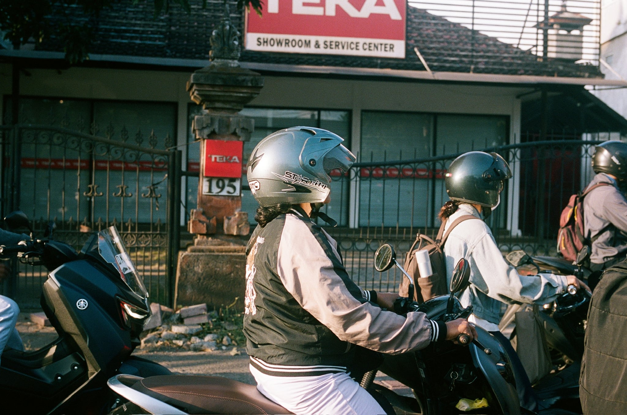 Helmeted scooter riders wait beside a roadside shopfront in afternoon light.
