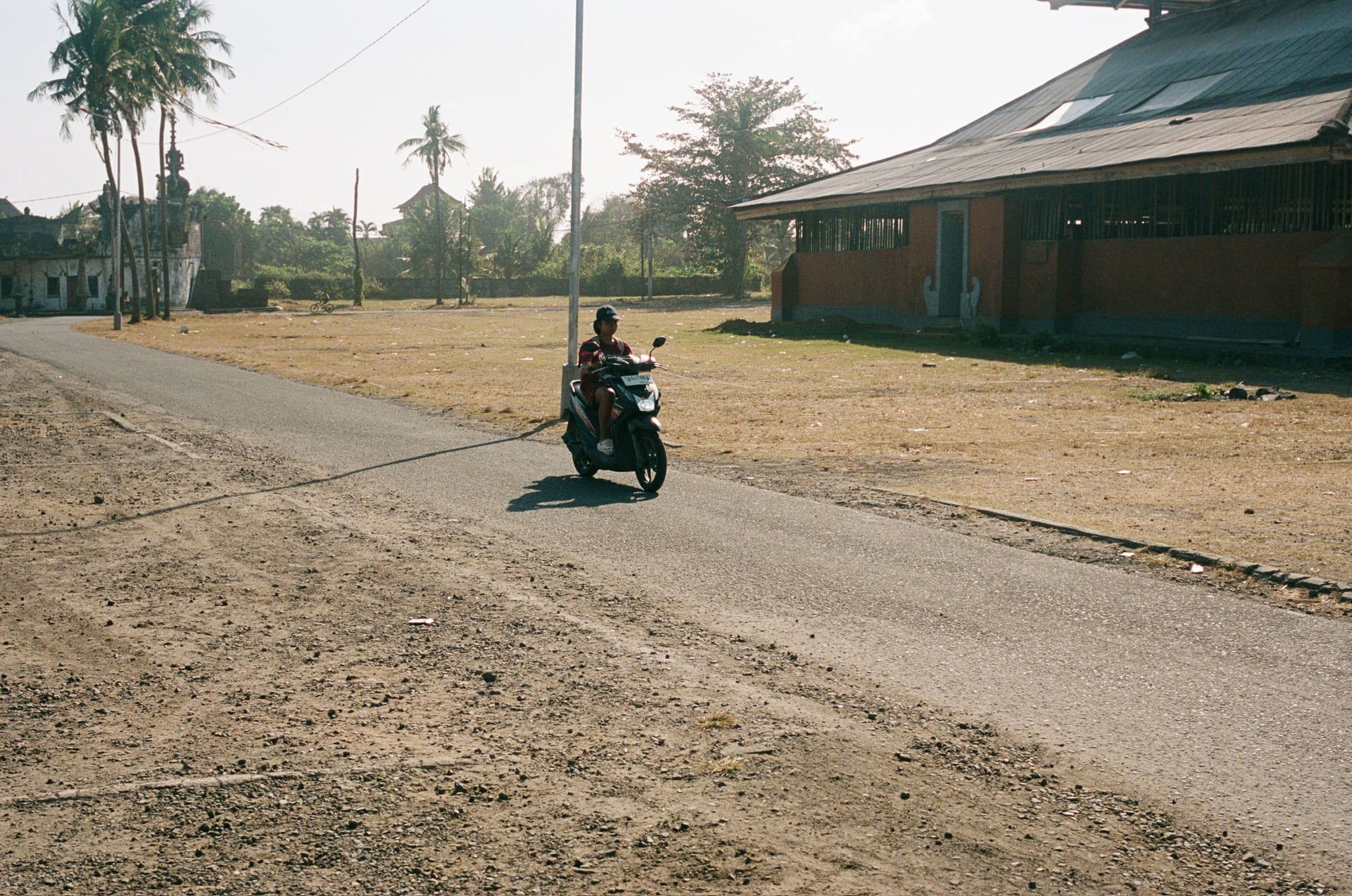 A scooter rider passes a broad tiered-roof pavilion beside open, dry ground.