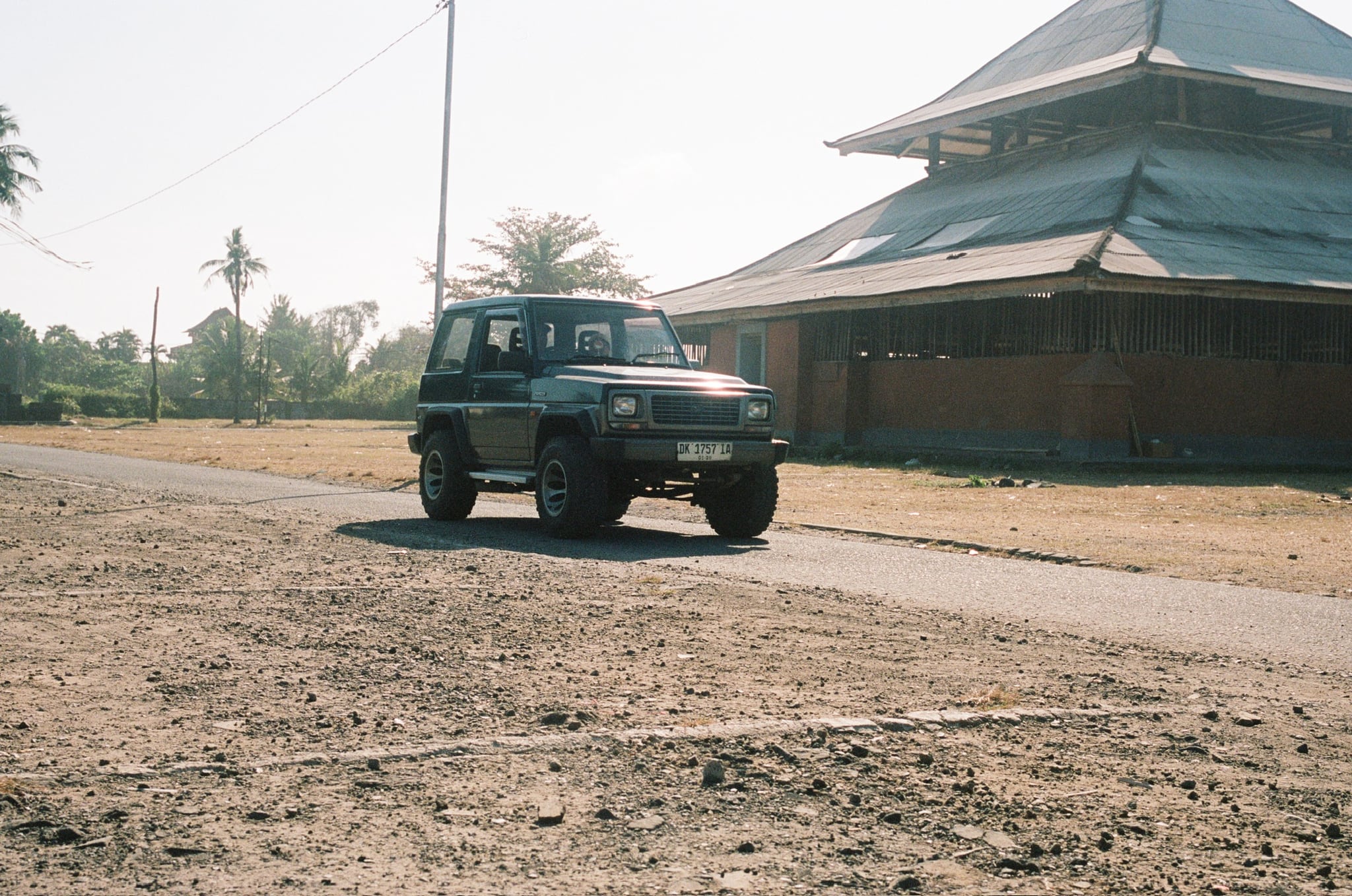 A dark off-road vehicle passes a large pavilion along a gravel road.