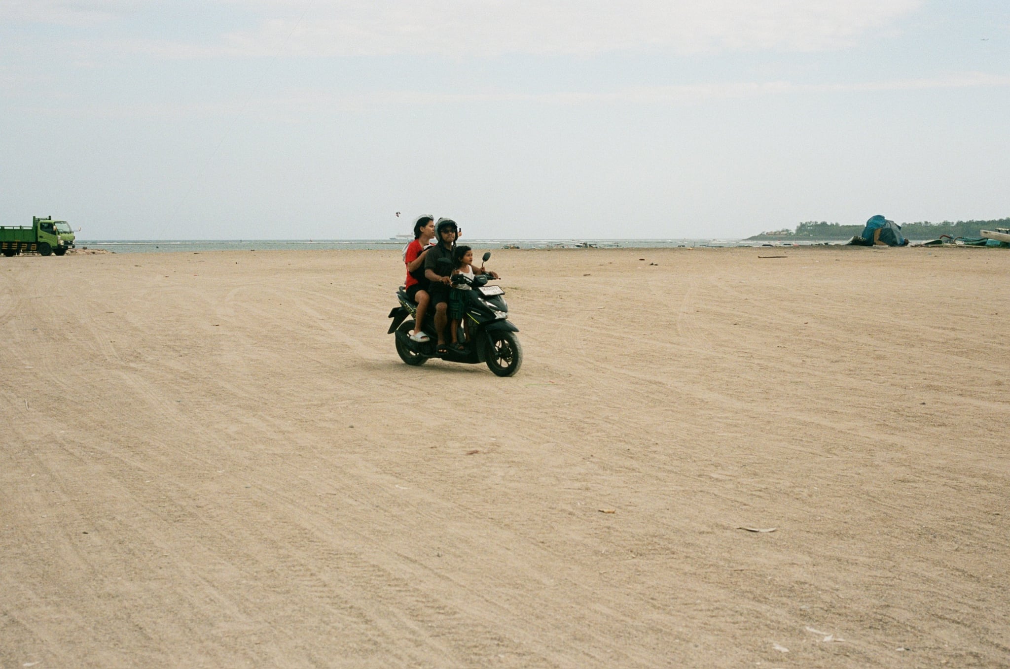 Two people ride a motorcycle across a wide, empty sandy field.