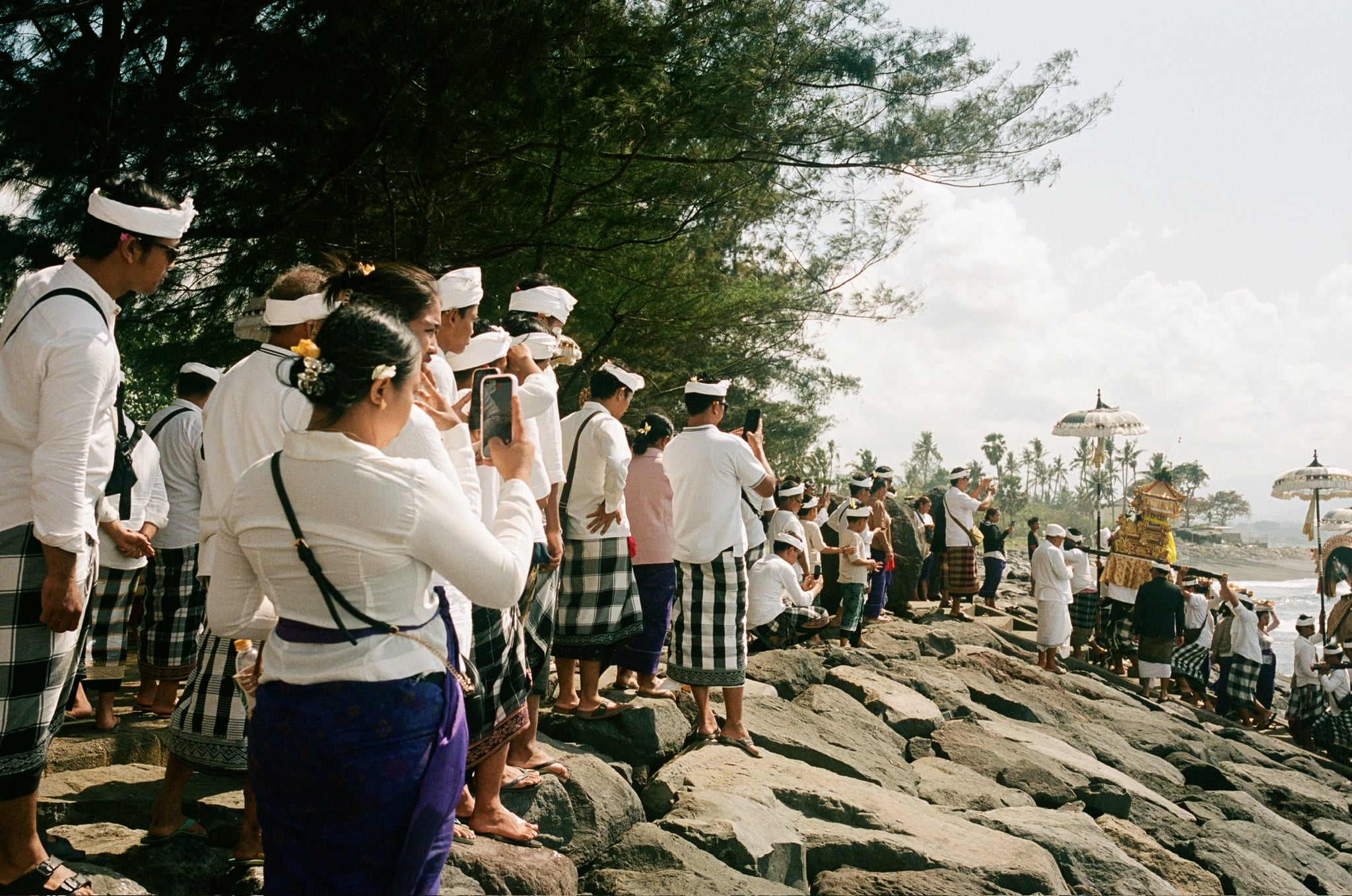 People stand along a stone embankment as one person raises a camera toward the coastal gathering.