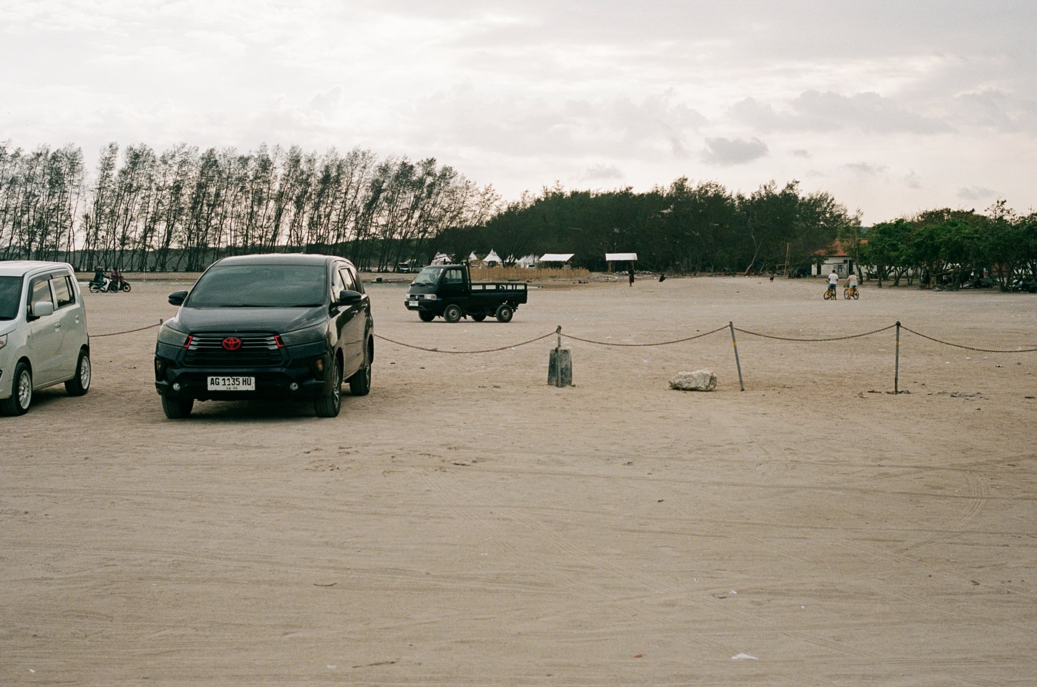Dark and pale vehicles cross a broad sandy lot bordered by trees.