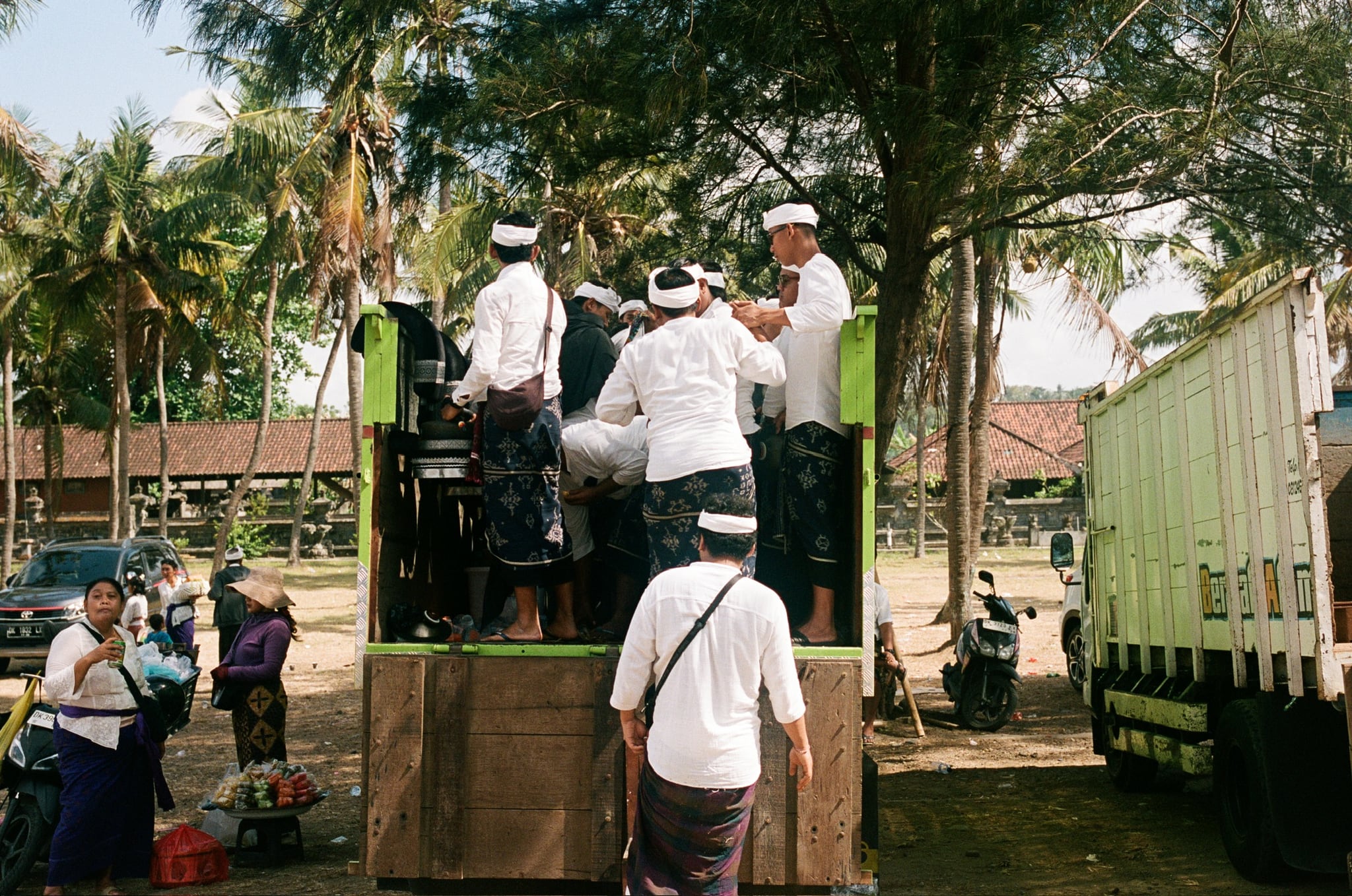 People in pale clothing stand in the back of a truck beneath trees, with a crowd nearby.