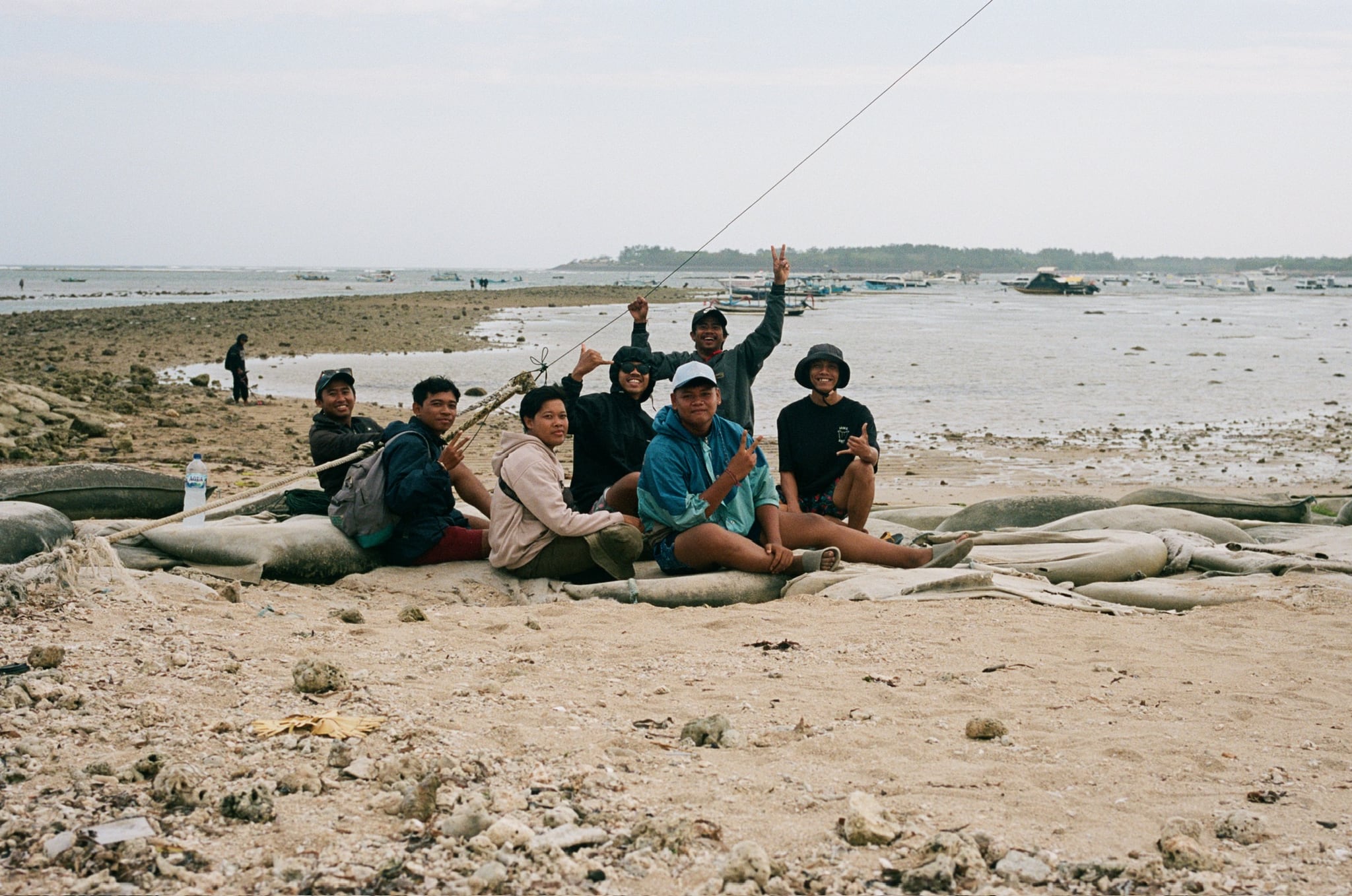 A group sits together on sandy ground near small boats and shallow coastal water.