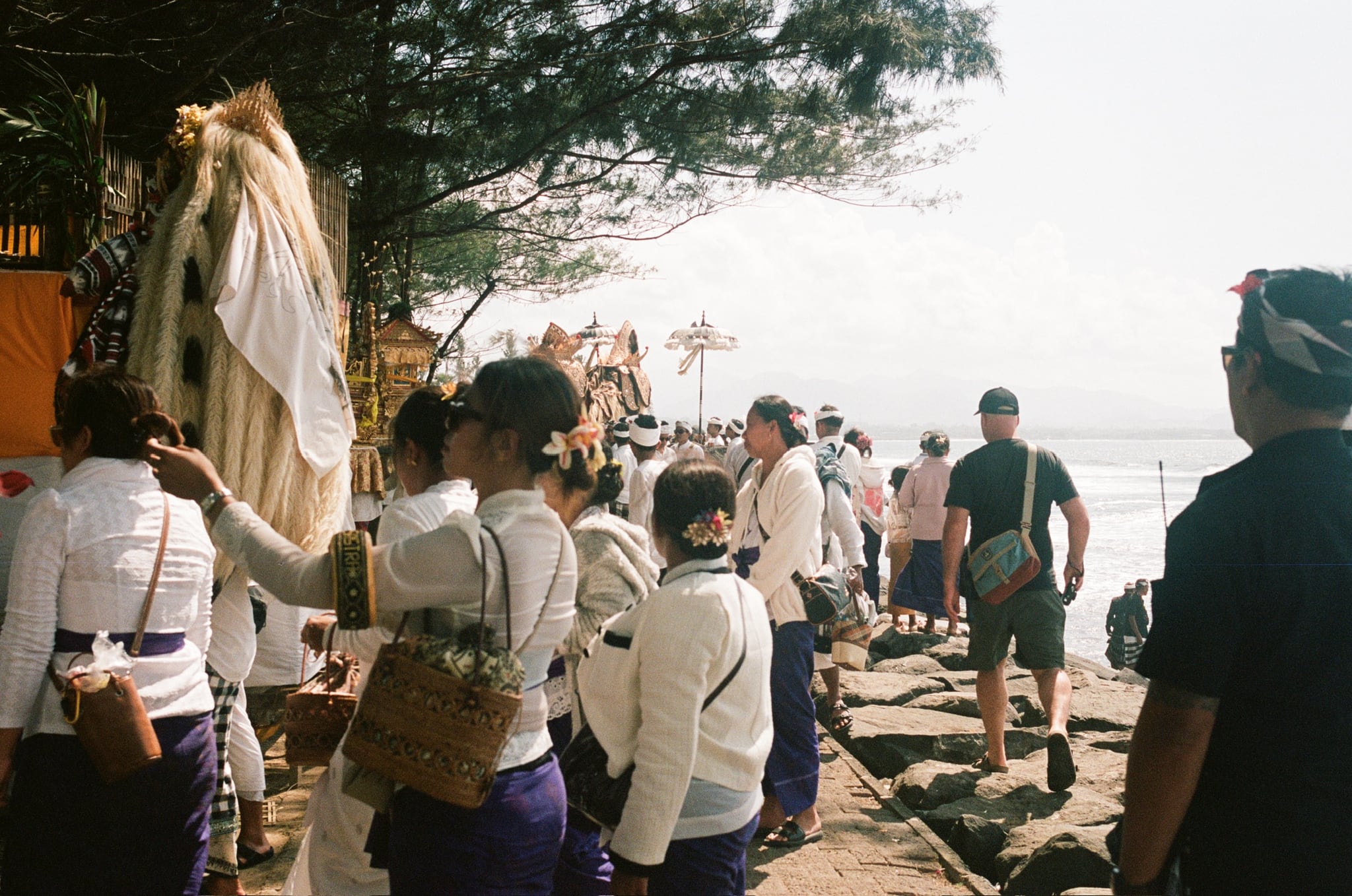 People in pale clothing gather beside trees and tall decorated objects near the sea.