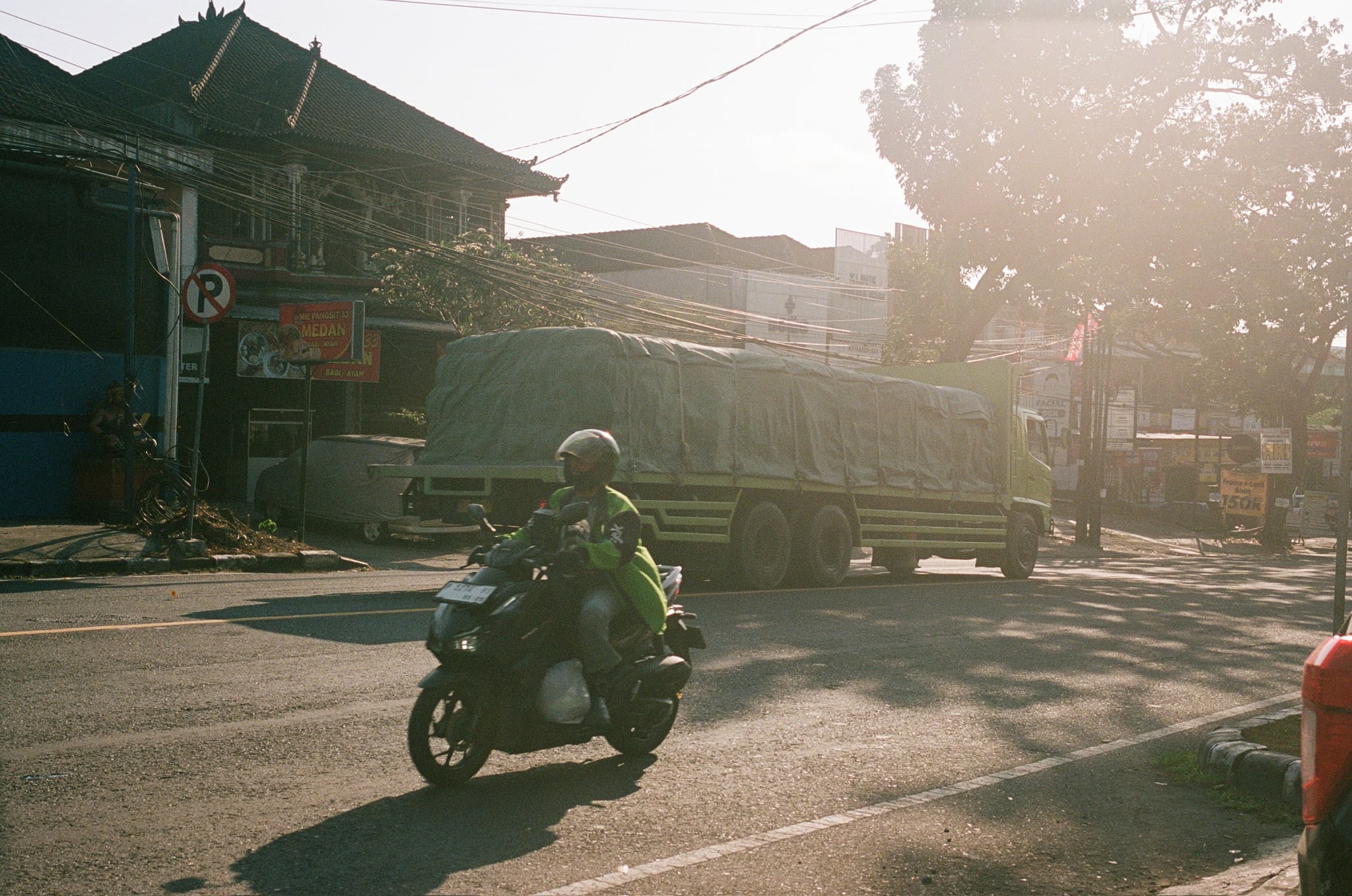 A scooter rider crosses a quiet street in warm, low sunlight beside dark shopfronts.