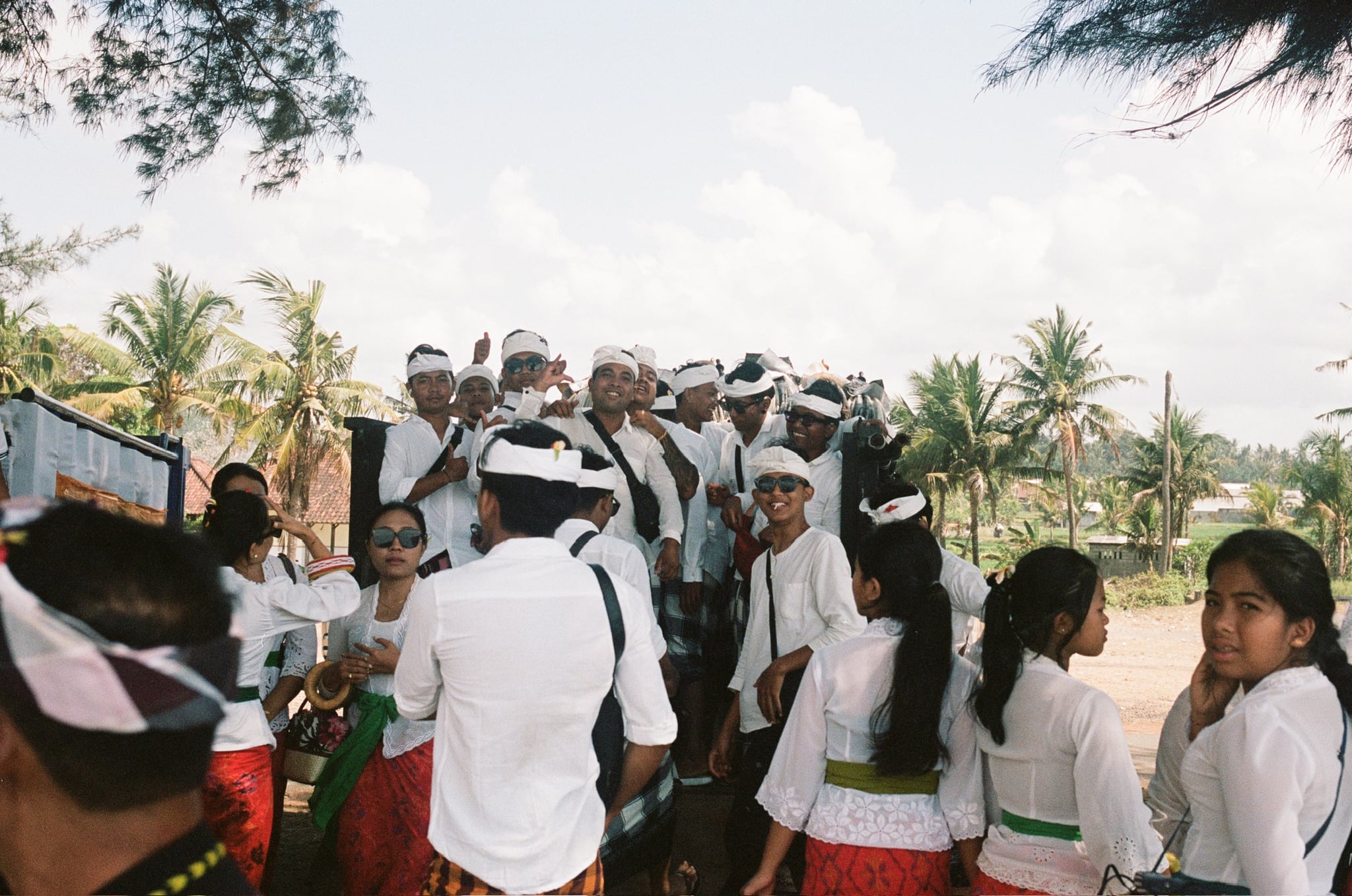 People in white and patterned clothing stand together beneath palms at an outdoor gathering.