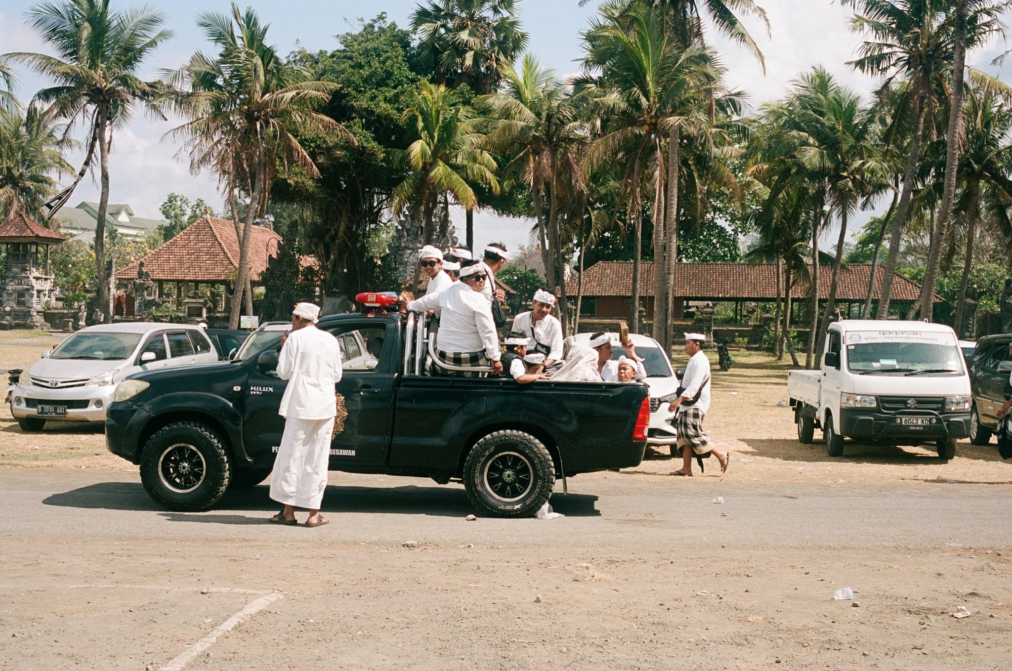People in white clothing ride in a dark pickup truck beneath palms beside an open lot.